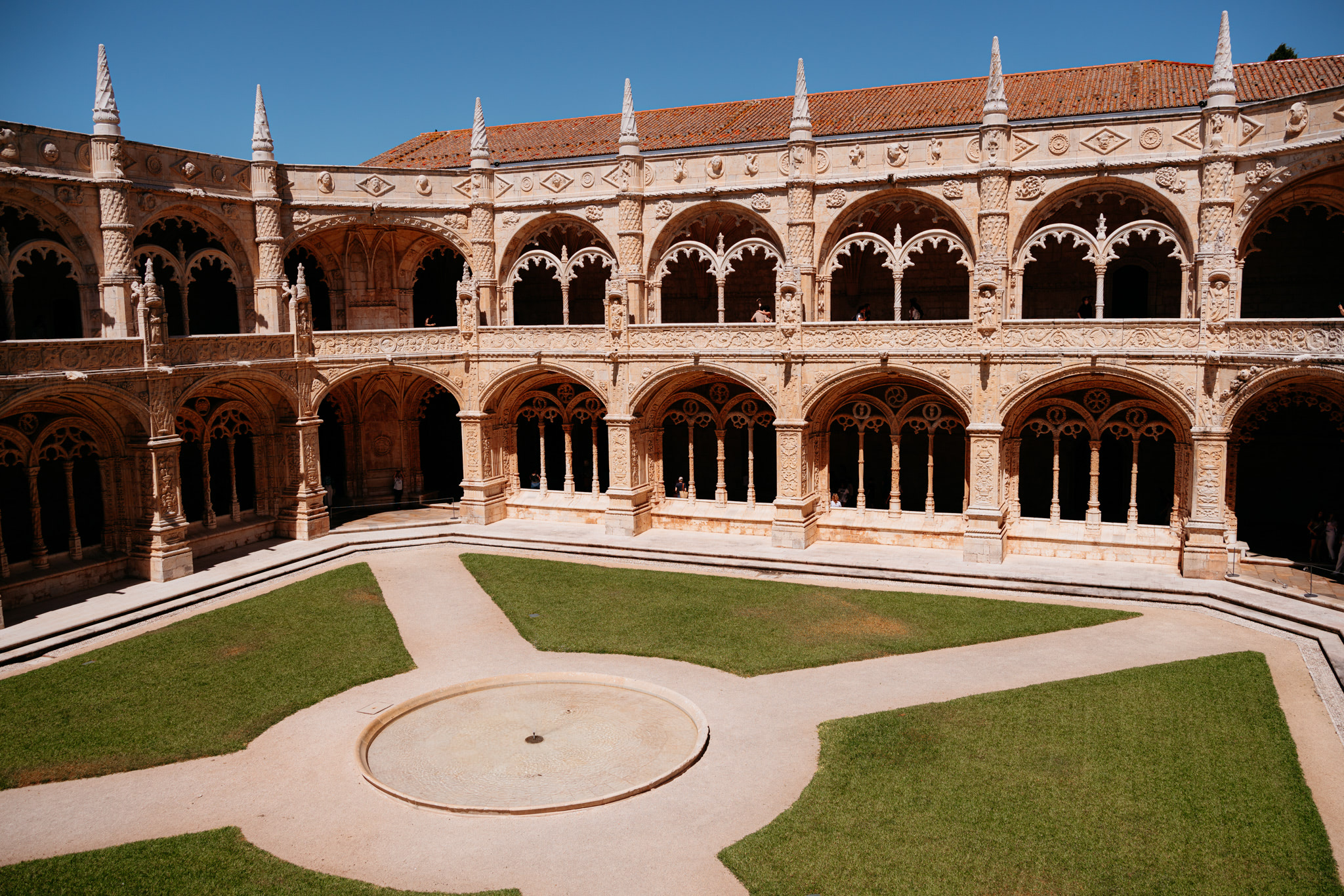 Jerónimos Monastery cloister with ornate arches, a central fountain, and a manicured lawn.