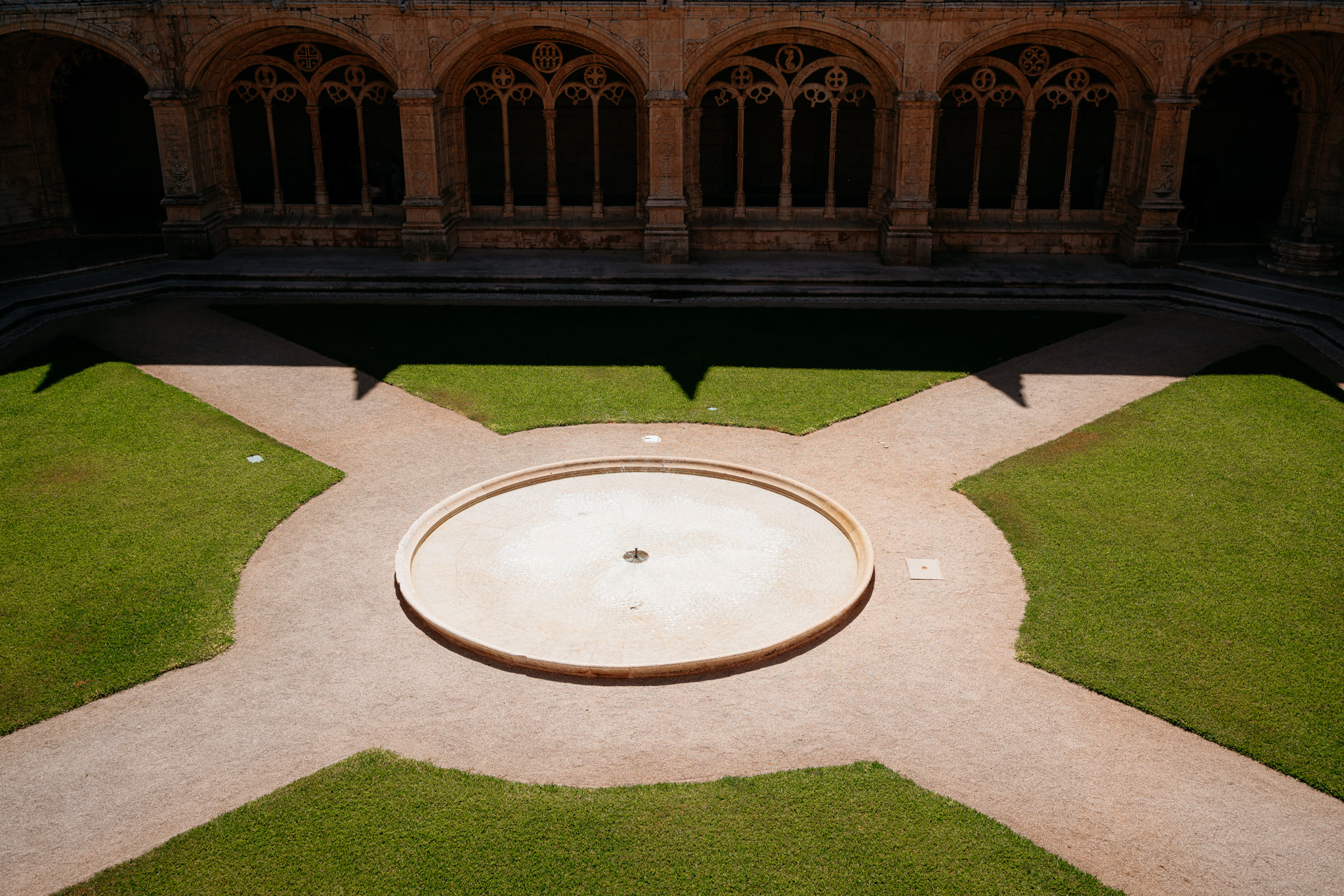 Overhead view of a courtyard with a circular fountain, gravel paths, and green grass, surrounded by a stone cloister.