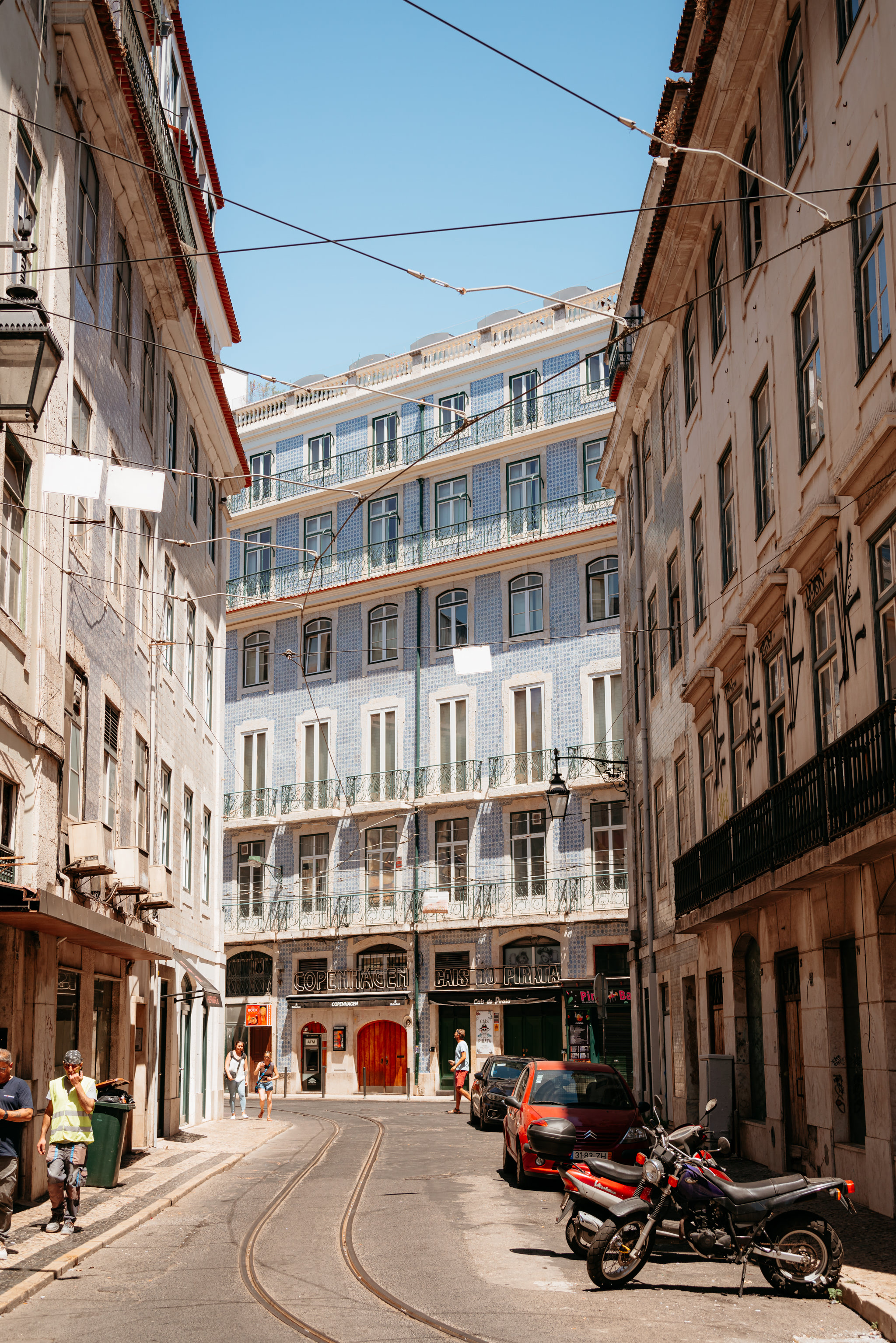 Narrow Lisbon street with blue tiled building, tram tracks, and parked vehicles.