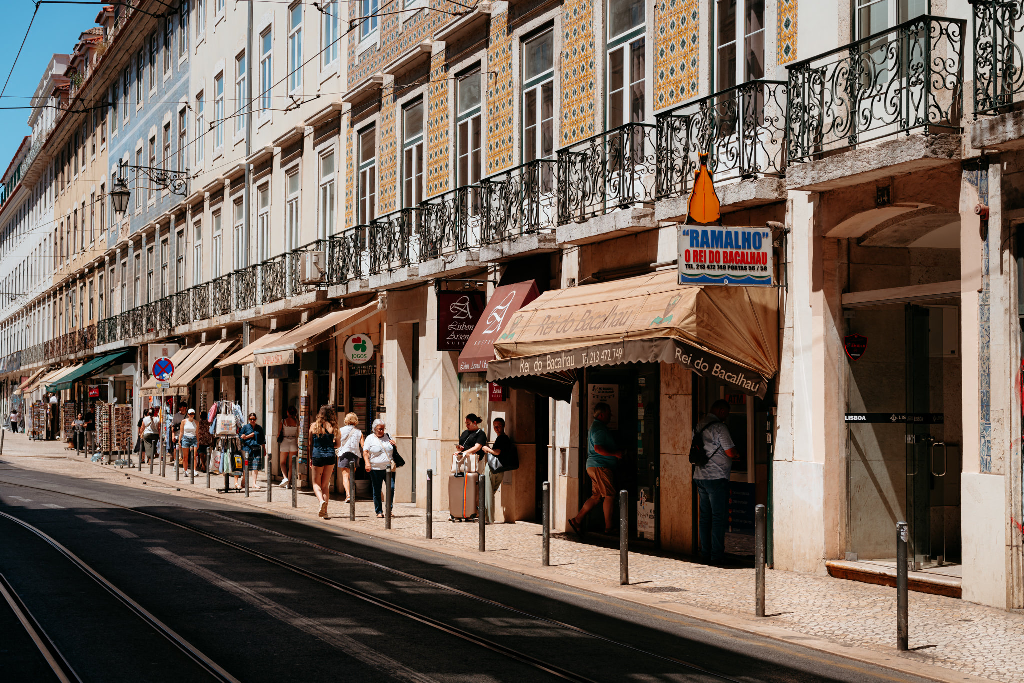 Sunny Lisbon street scene with pedestrians and shops, featuring ornate balconies and tiled buildings.