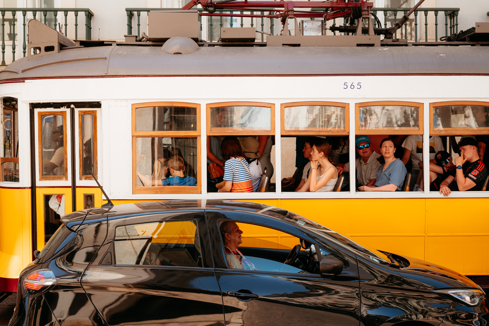 A yellow and white Lisbon tram, number 565, full of passengers, next to a black car with a woman driver.