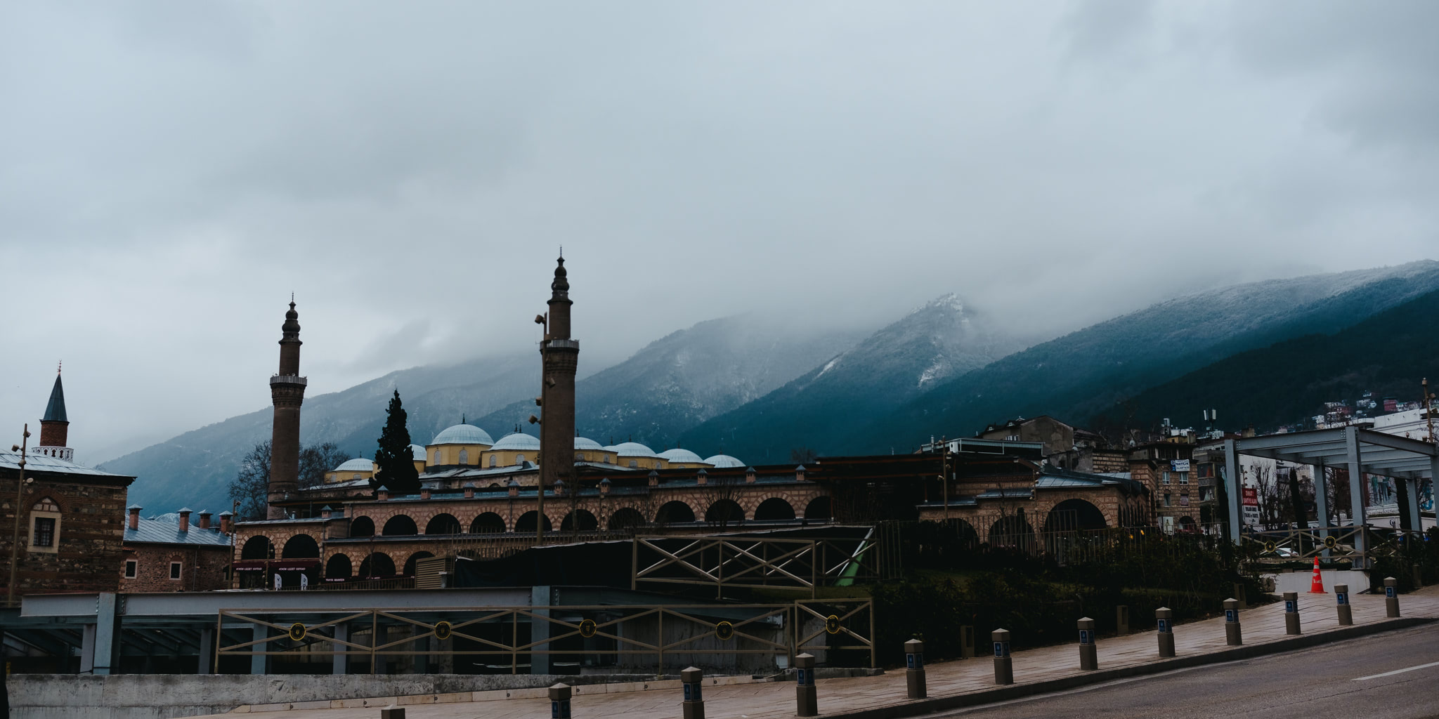 Historic mosque with minarets and domed roofs against a backdrop of mountains under an overcast sky.