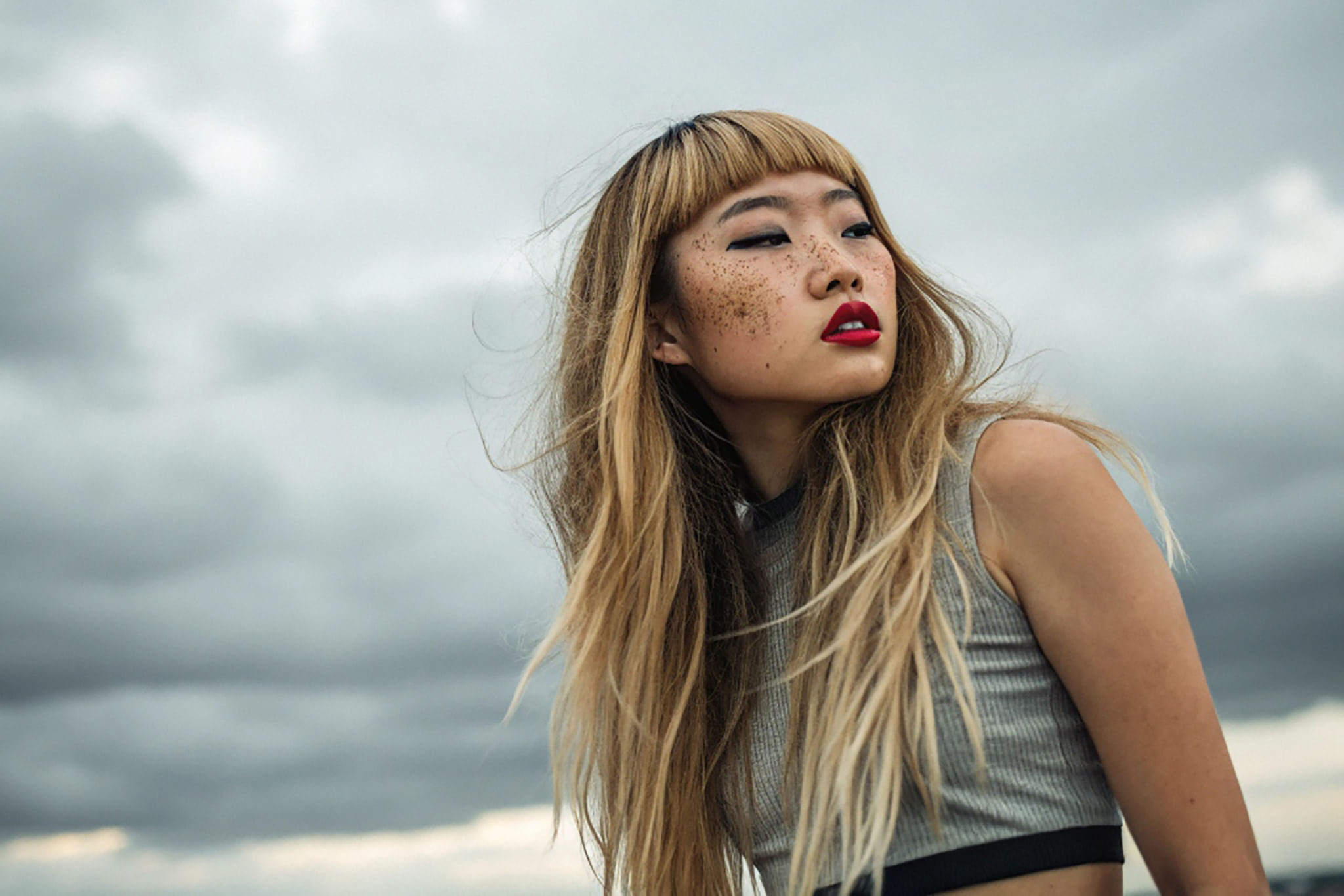 Portrait of a woman with long blond hair and freckles, looking off to the side against a cloudy sky.