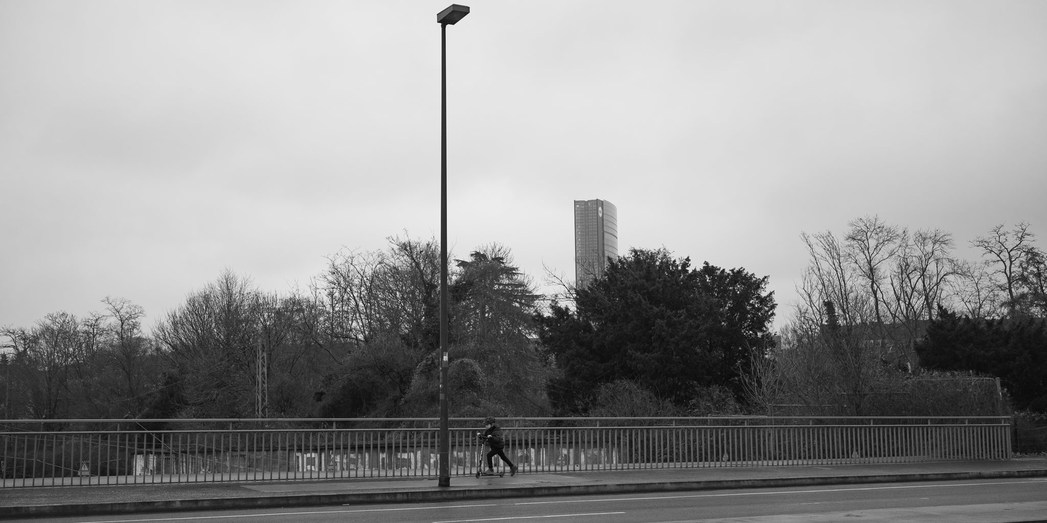 A monochrome shot of a boy on a scooter on a bridge, with trees and a tall building in the background.