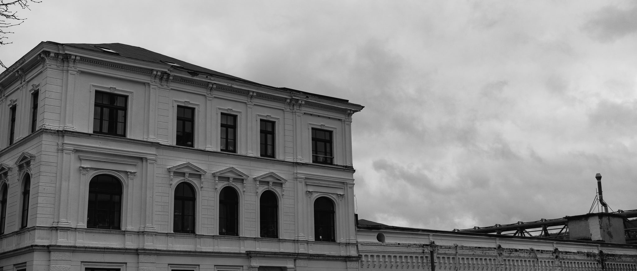 Black and white photo of a building facade with arched windows, rectangular windows, and decorative molding against a cloudy sky.