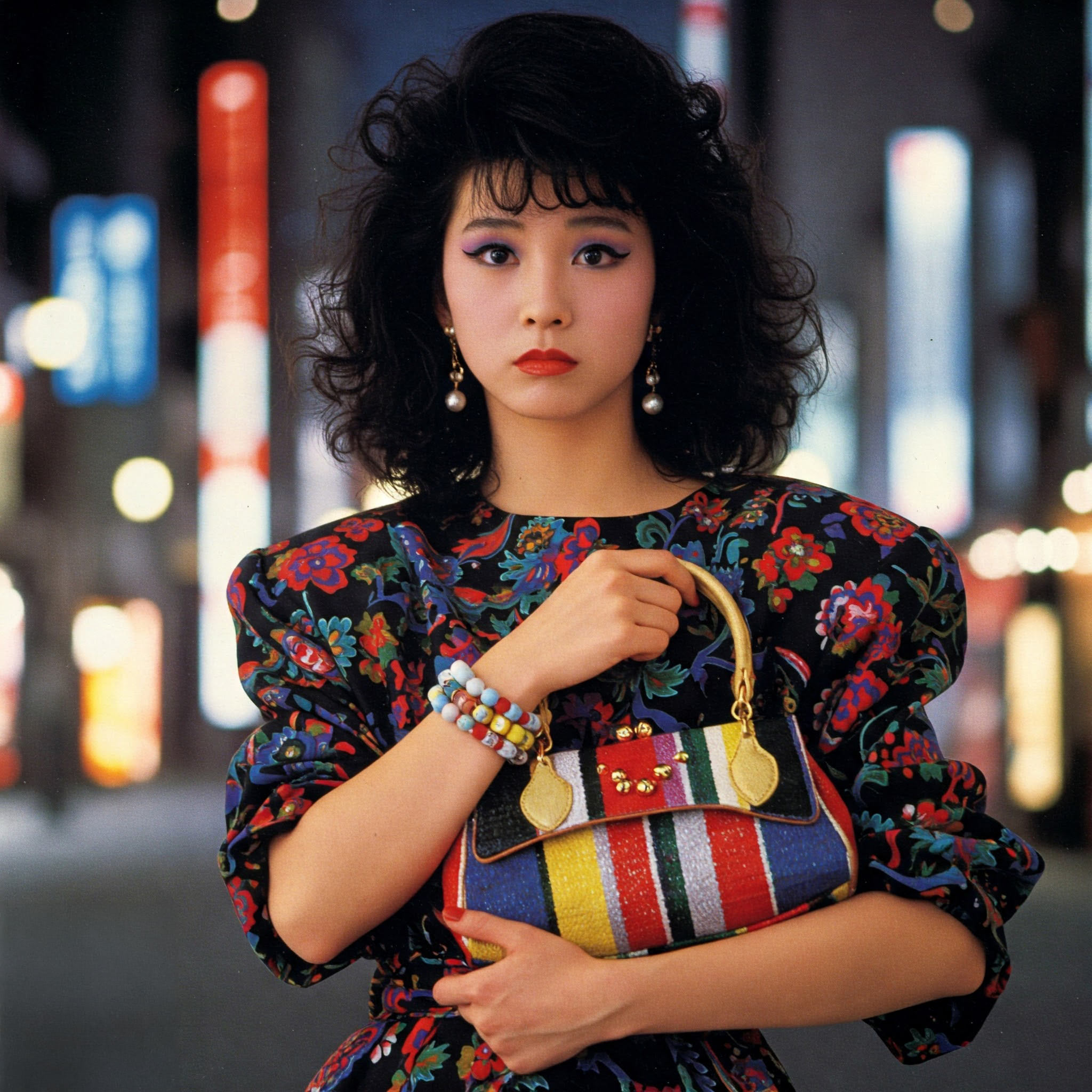A woman with dark hair and colorful makeup holding a striped handbag, with city lights in the background.