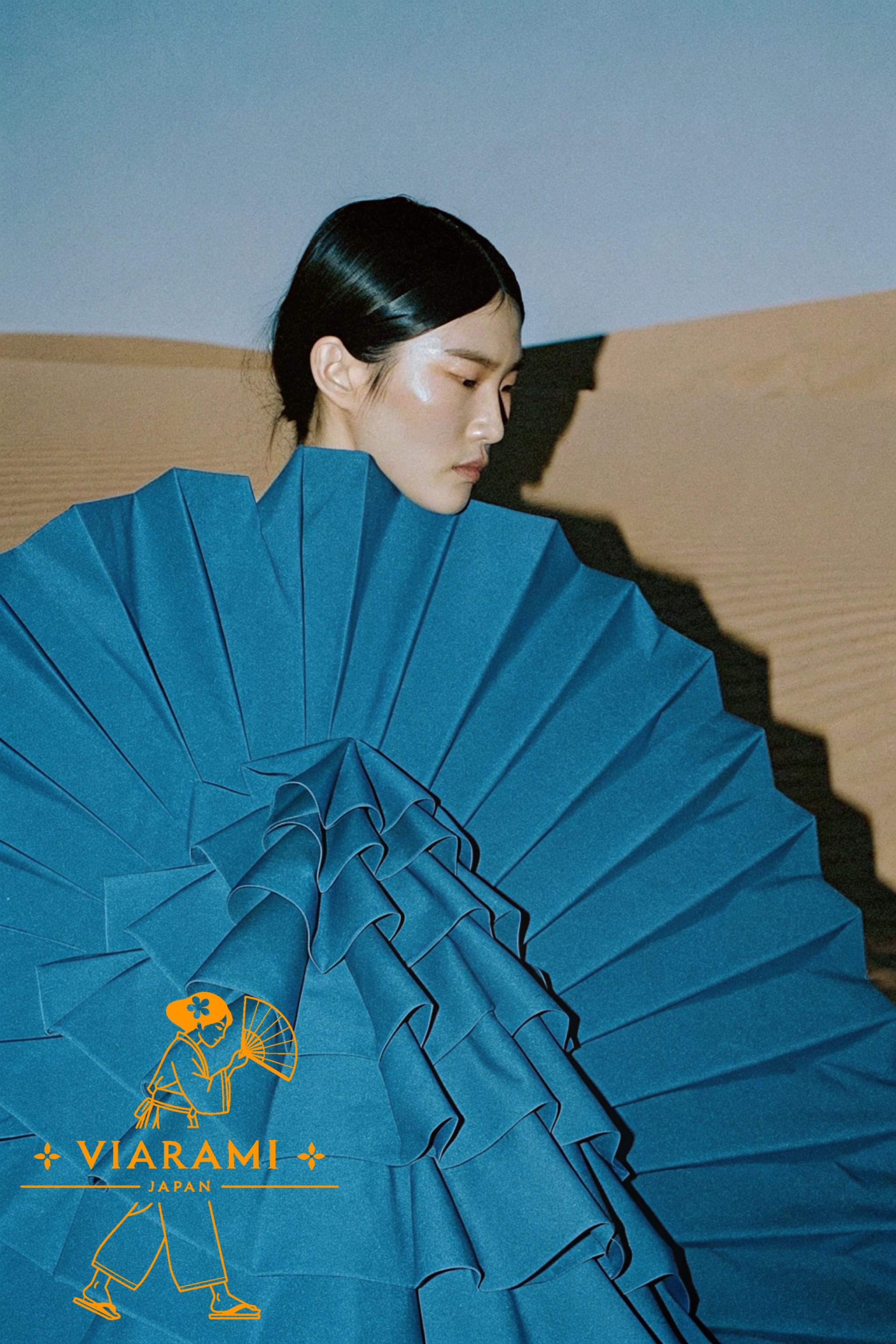 Asian model wearing a large, blue, origami-style dress in a desert setting.
