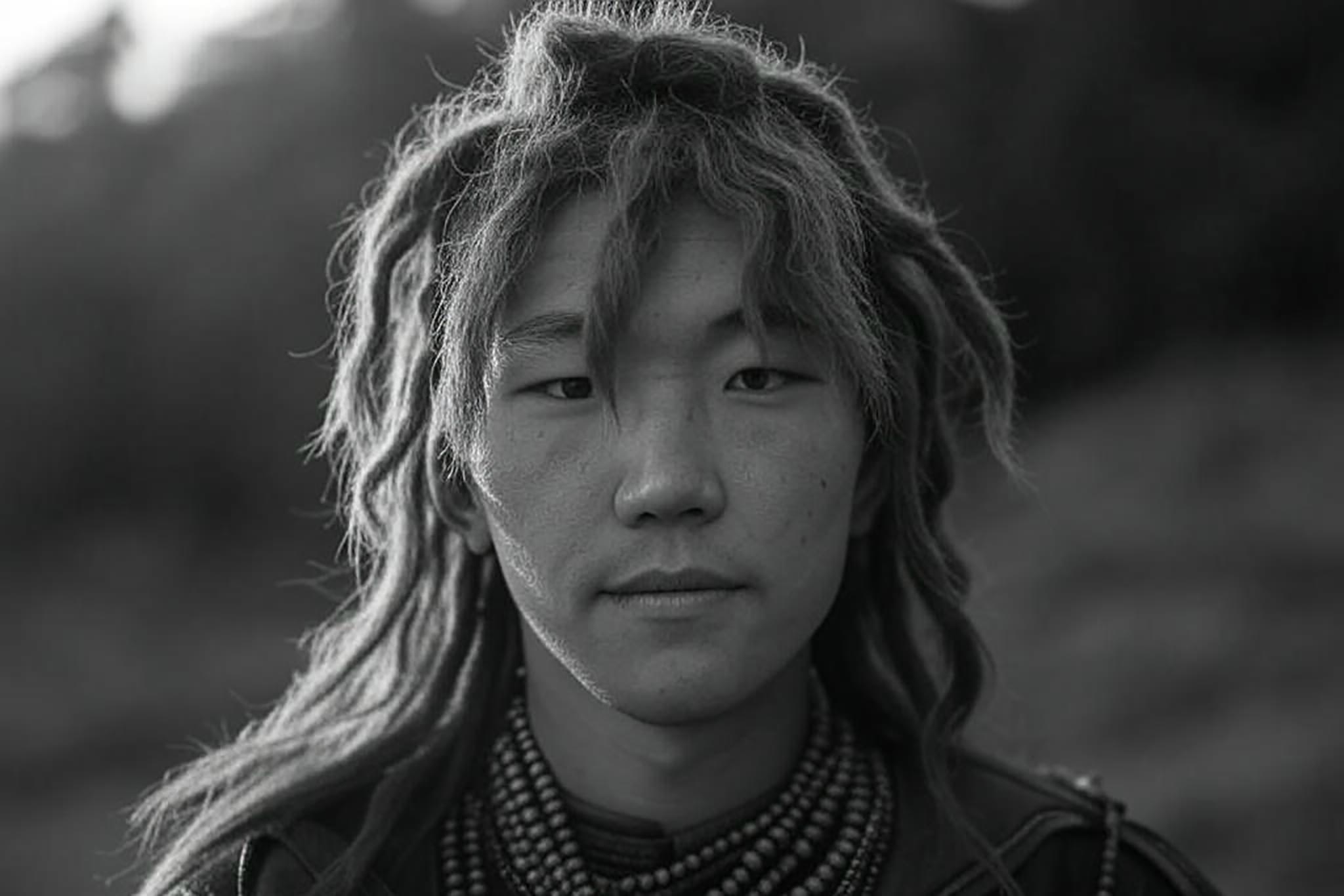 Black and white portrait of a young man with long hair and beaded necklaces.