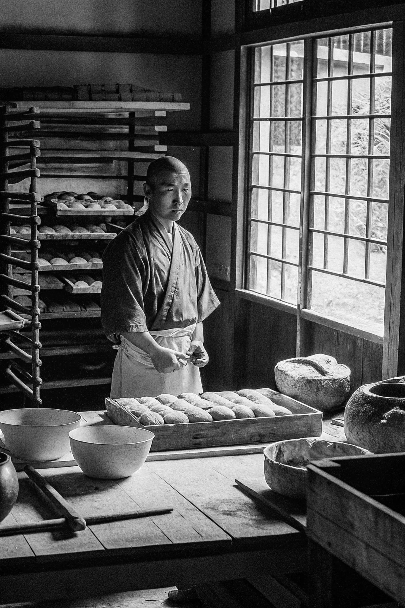A baker standing next to a window, wearing an apron and making bread.