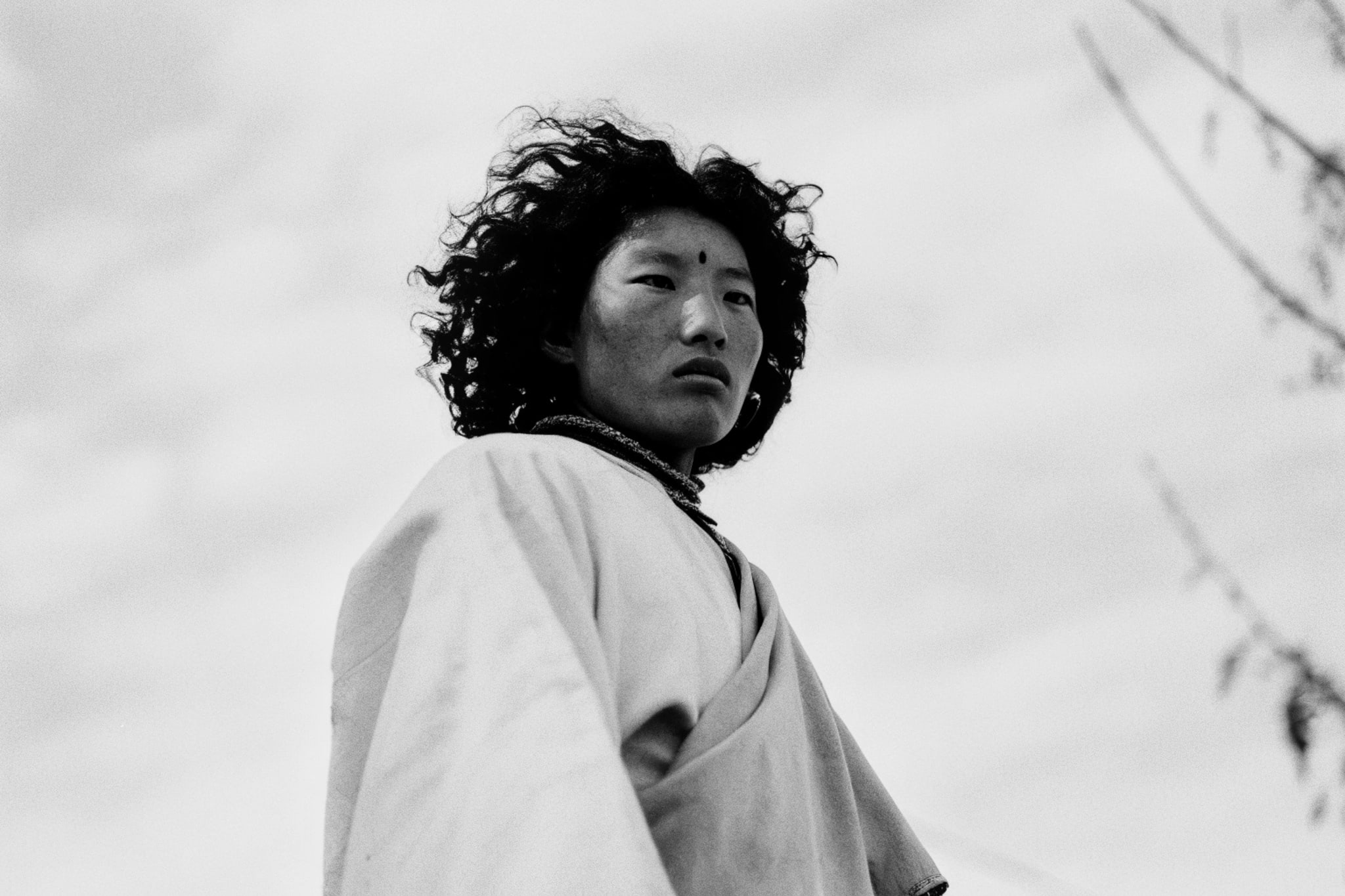 Black and white portrait of a person with dark curly hair, wearing a robe and necklace, looking upward.