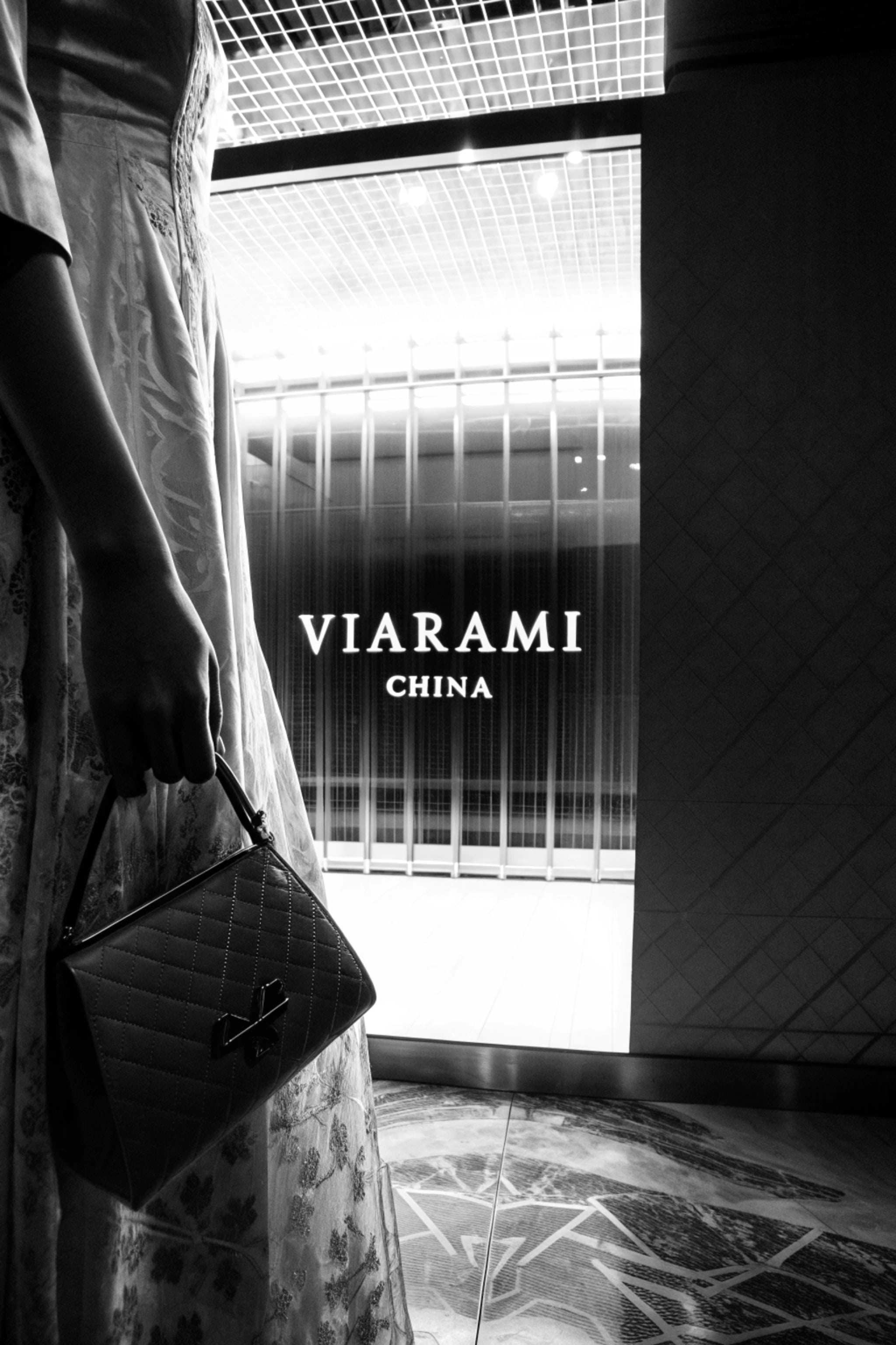Black and white photo of a woman in a patterned dress holding a handbag in front of a storefront.