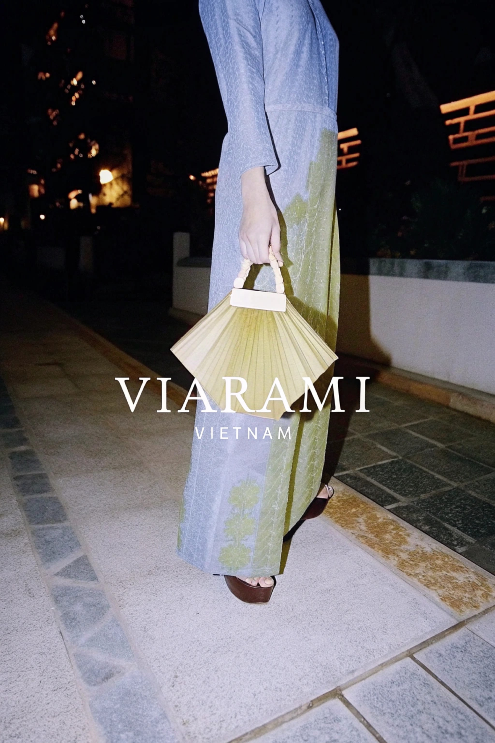 A person in a blue and green dress holds a tan fan-shaped purse at night.