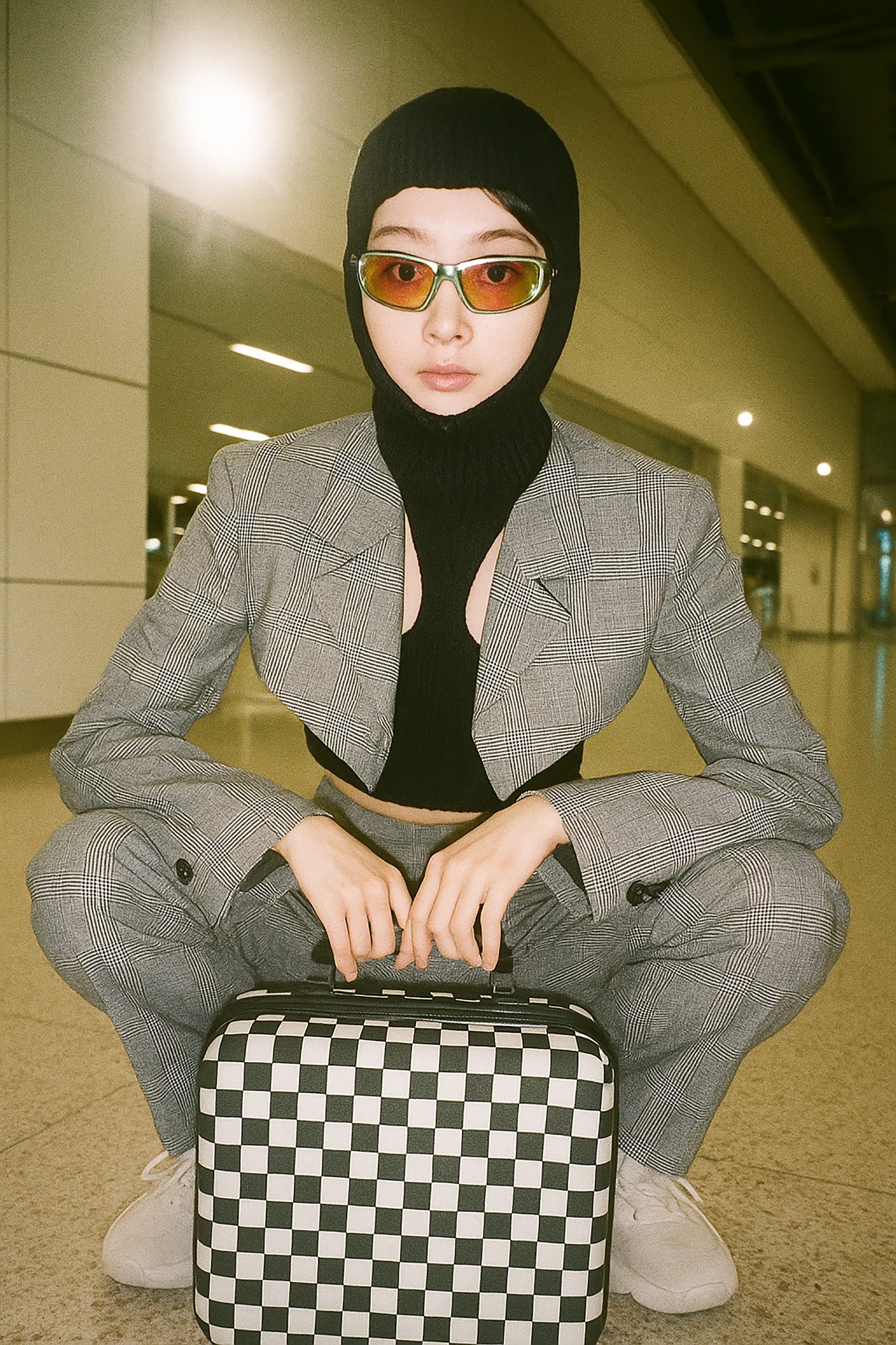 A woman in a balaclava, sunglasses, and a checked suit squats with a black and white checkered suitcase.