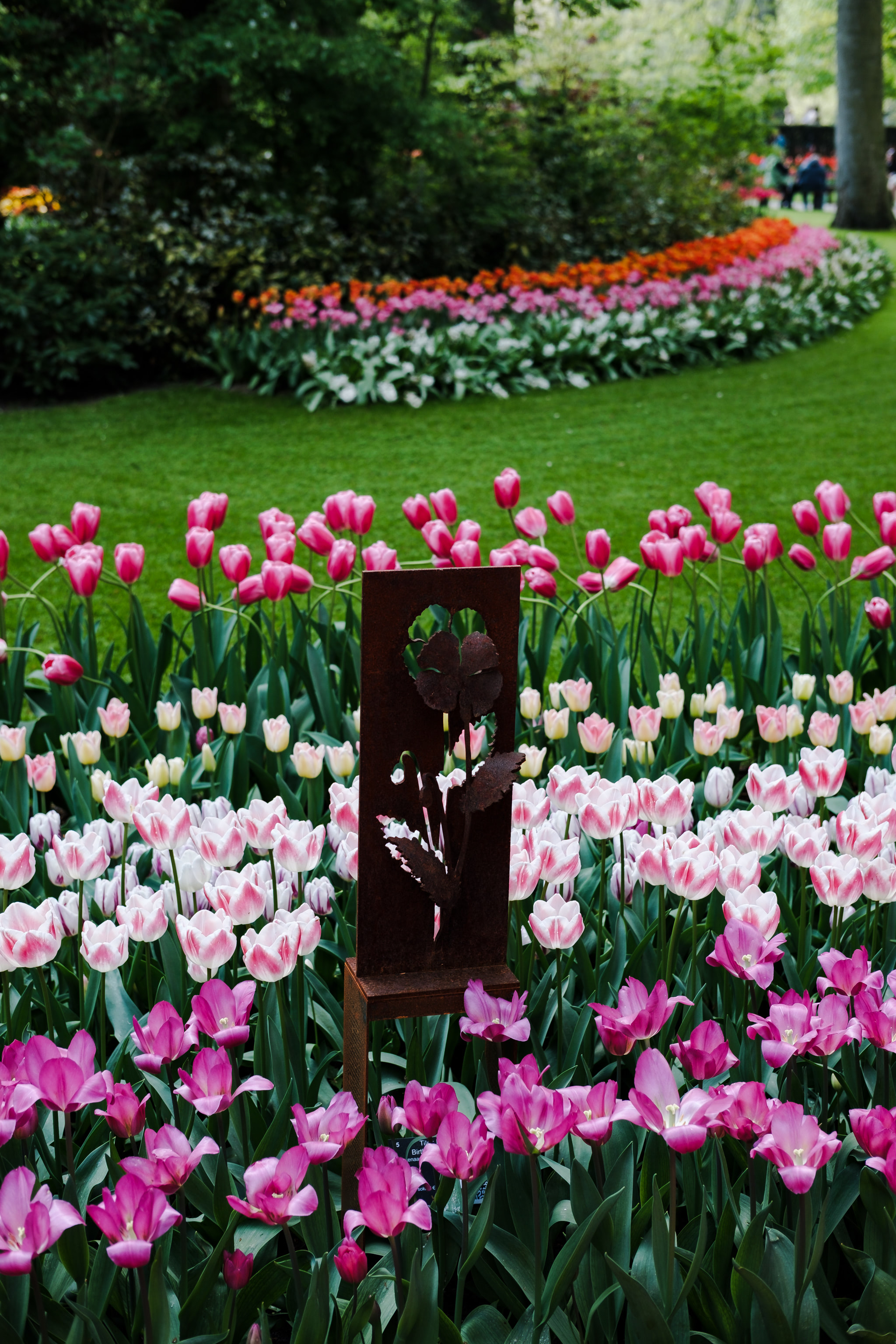 A garden filled with pink, white, and purple tulips. A metal cutout of a flower stands in the center of the foreground. A backdrop of green shrubs.