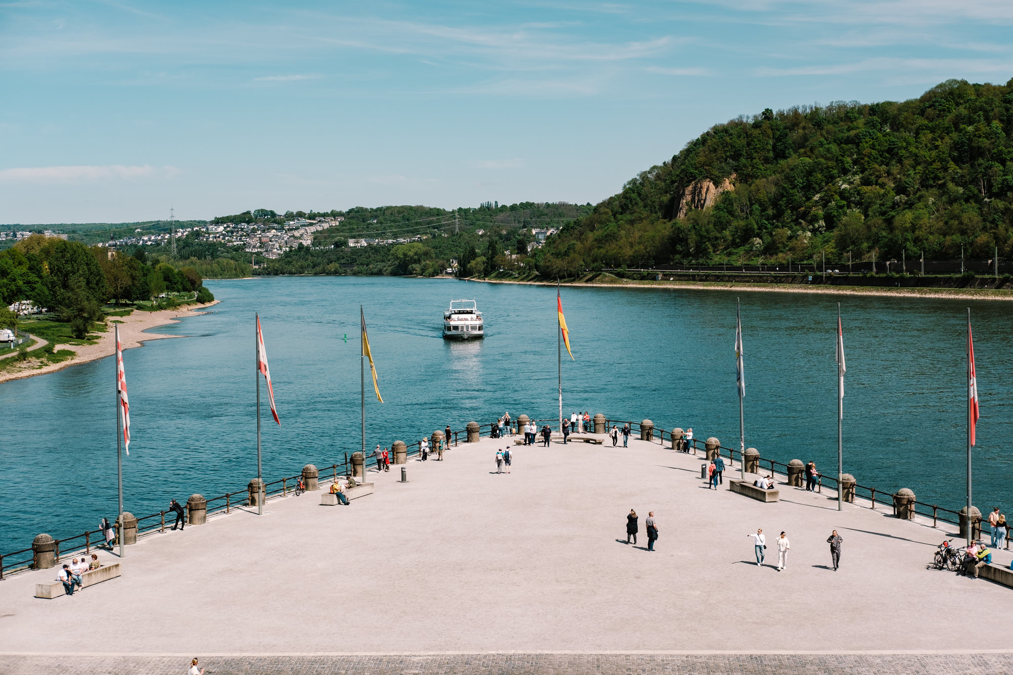 People on a waterfront terrace overlook a wide river with a boat. Flags line the terrace. Hills and buildings are visible in the distance.