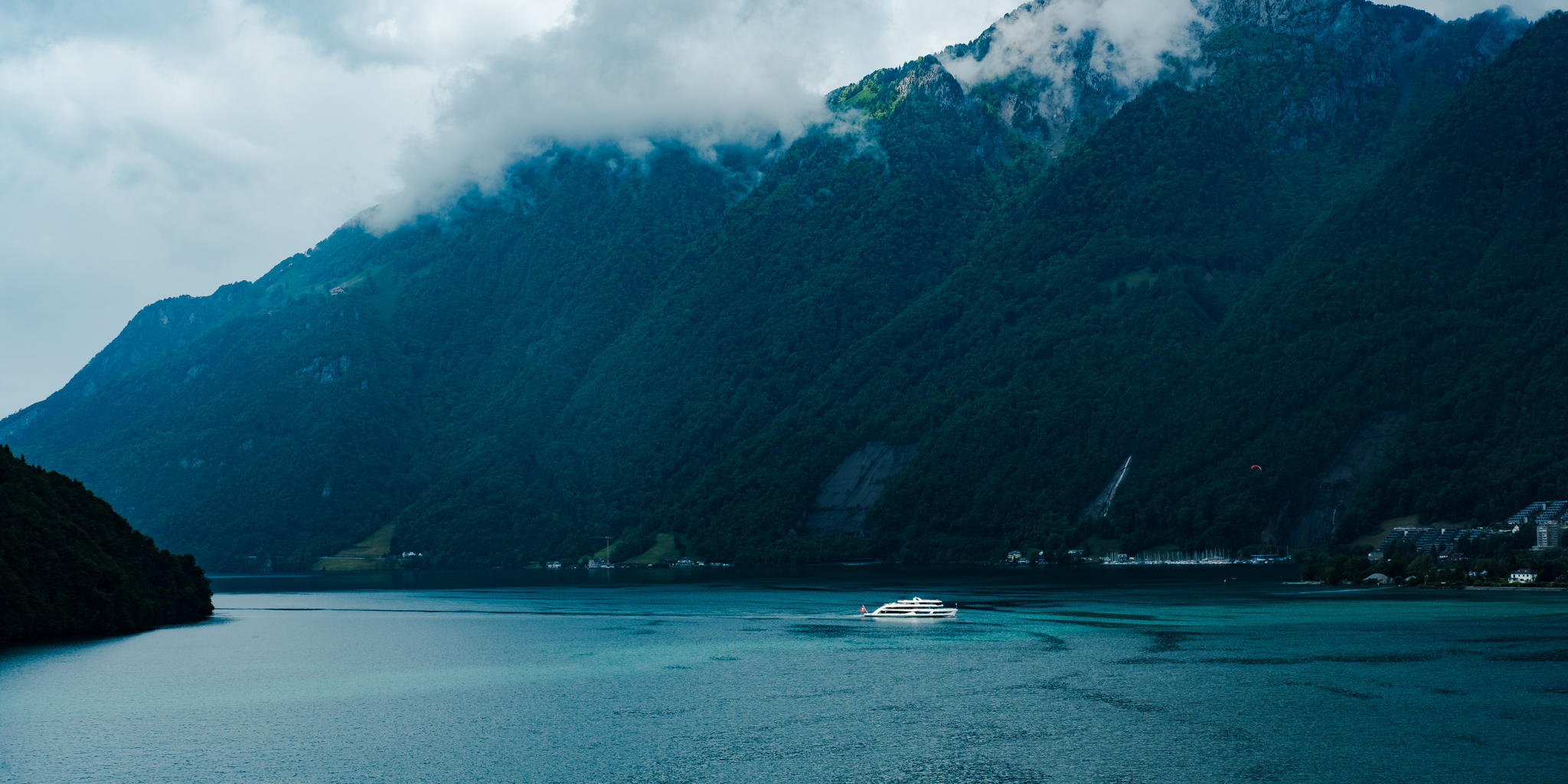 Lake with a white boat and mountains covered in trees, partially shrouded in clouds.