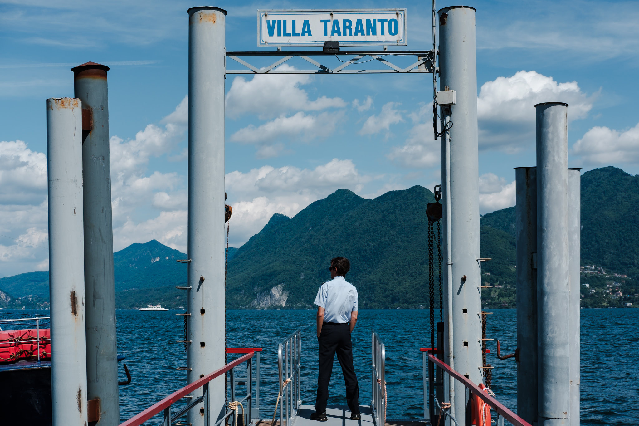 A person standing on a pier with a sign that says 'VILLA TARANTO'. Blue water and green mountains in the background.
