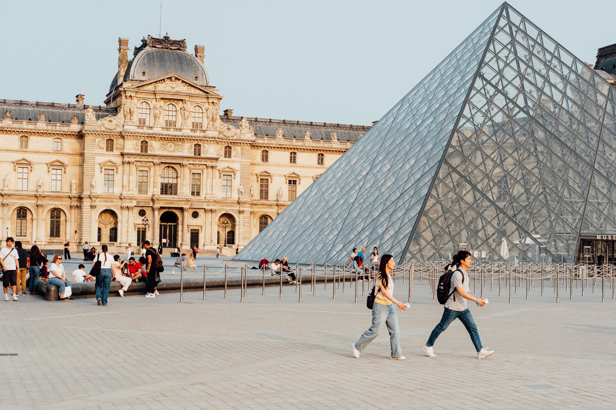 Exterior view of a modern glass pyramid and ornate building with people walking.