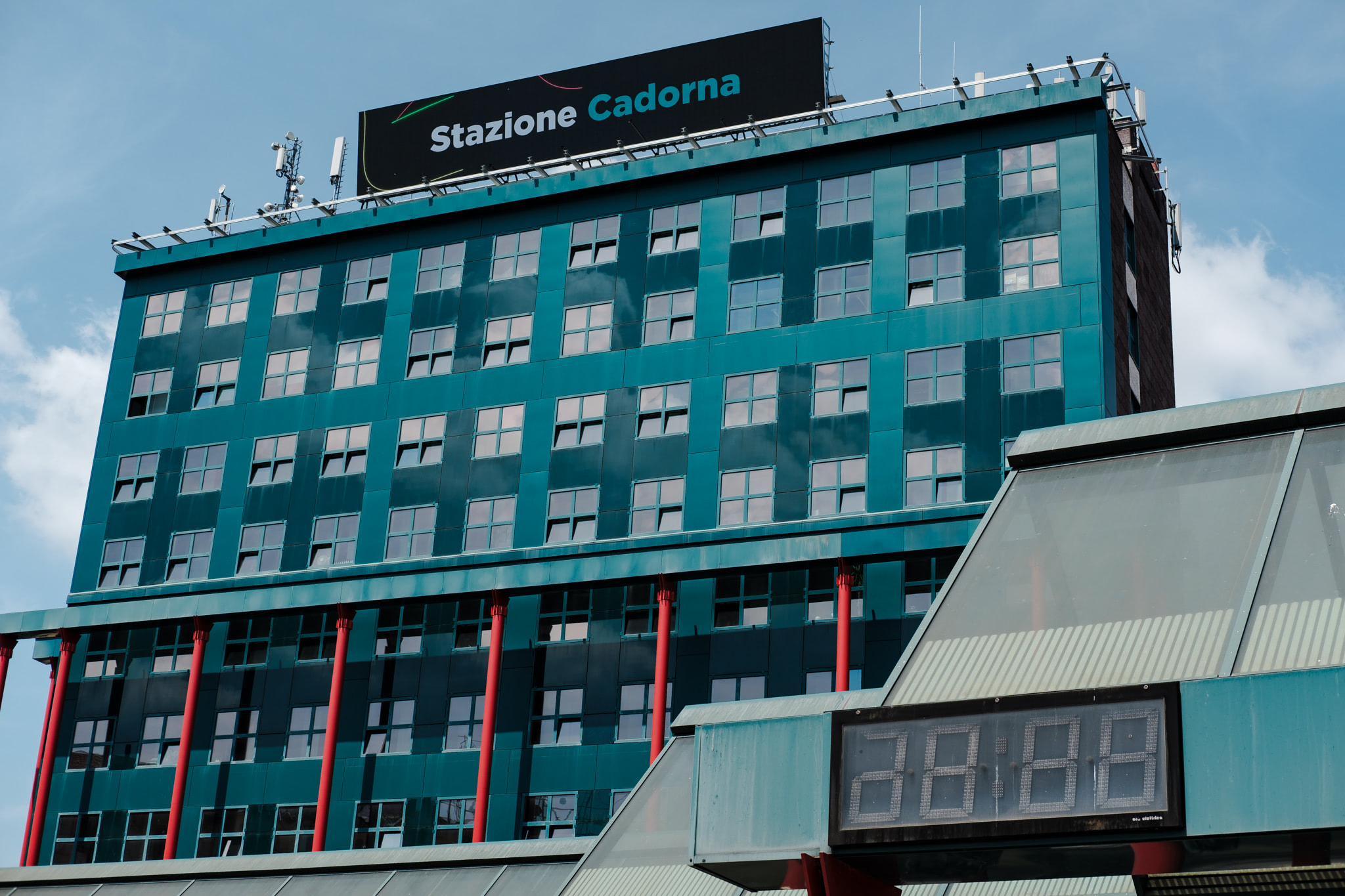 Teal building with numerous windows, red pillars, and a digital clock against a blue sky.