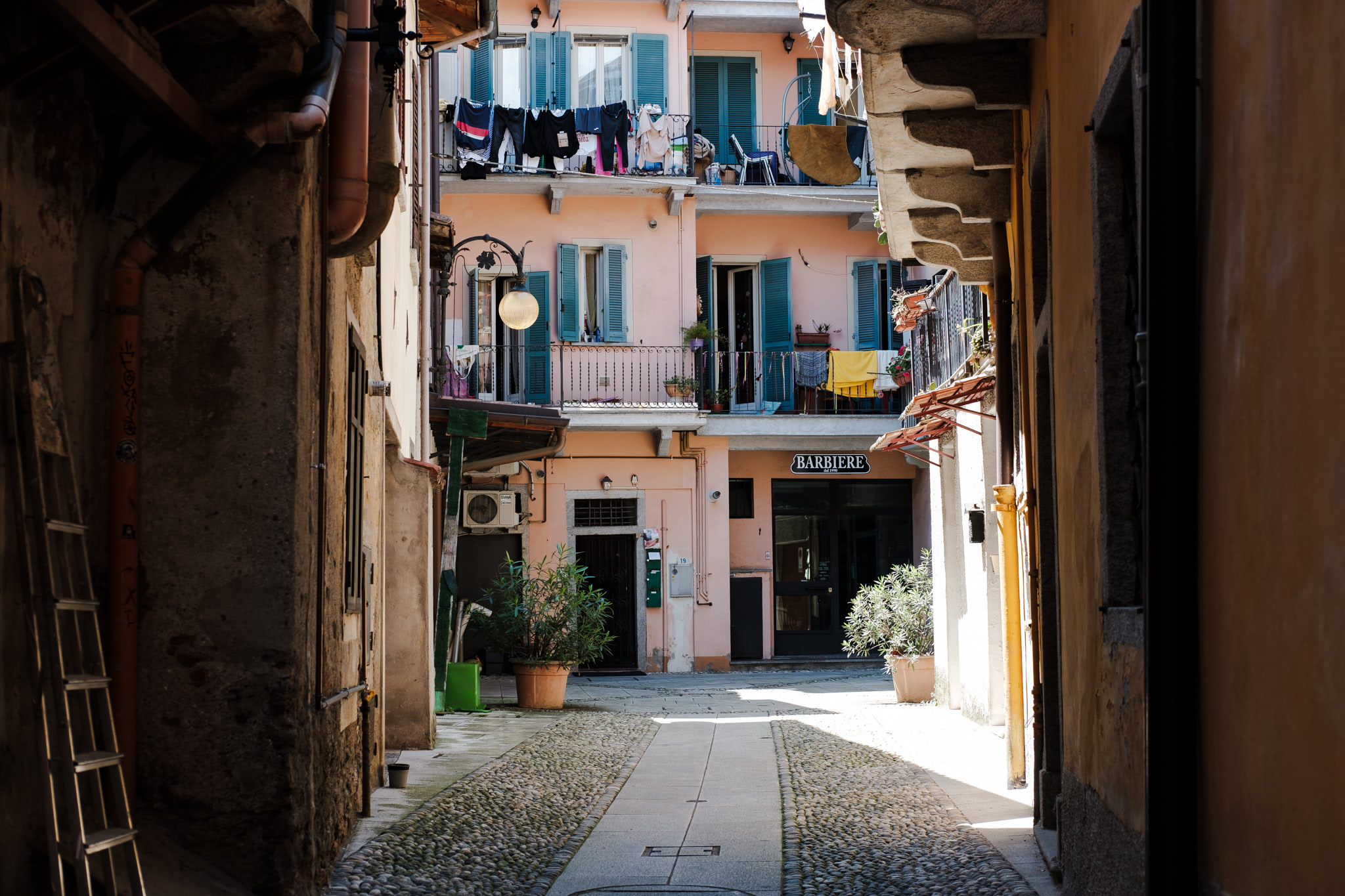 Narrow cobblestone street in an Italian town, framed by buildings. Laundry hangs on balconies of a pink building. A barbershop sign is visible.