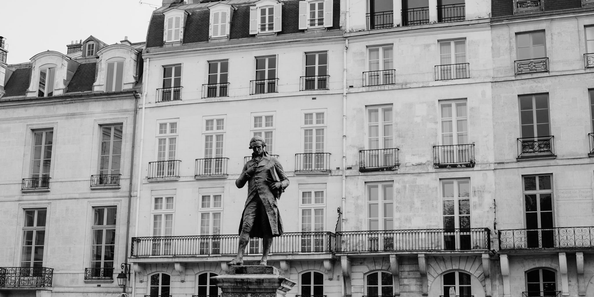 Black and white image of a statue in front of a building with windows, balconies, and architectural details.