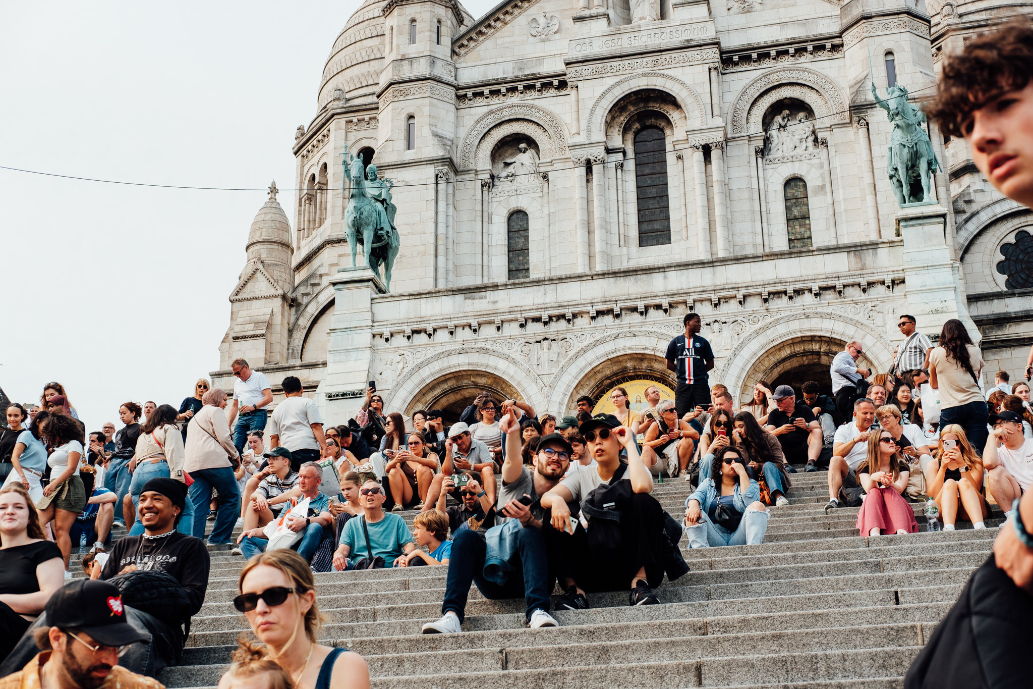 People sitting on steps in front of a large church. A statue of a man on a horse is visible.
