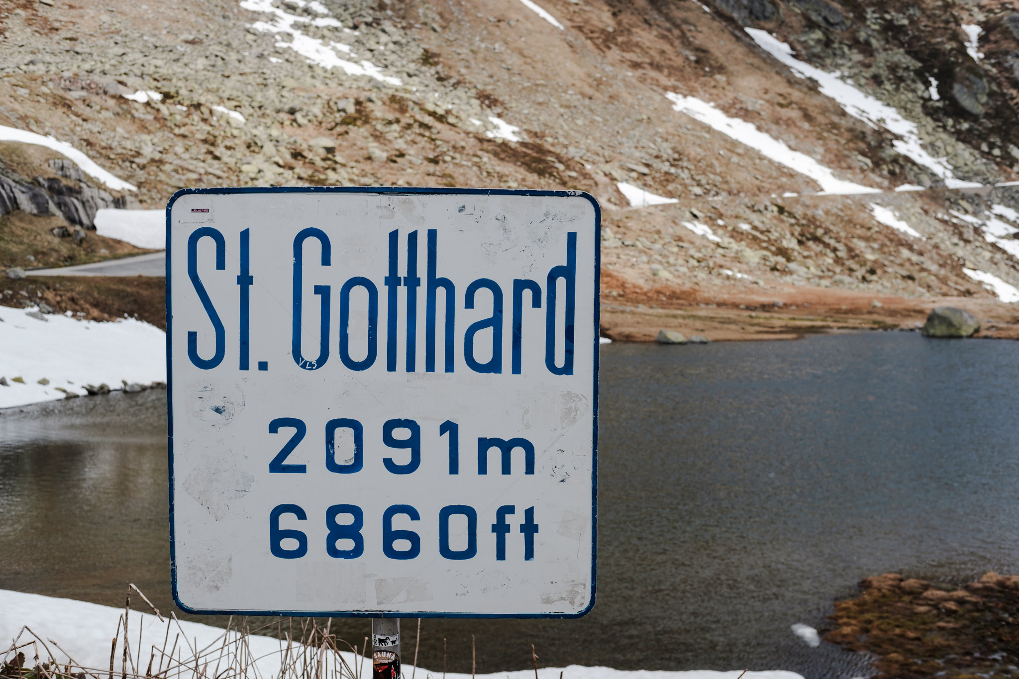 Sign for St. Gotthard with altitude information (2091m, 6860ft), mountains and lake in background.
