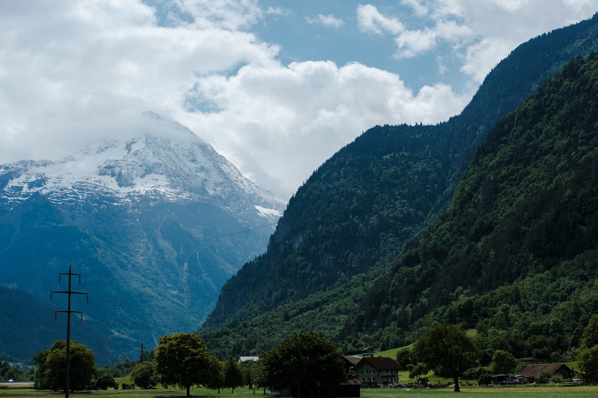 Mountain landscape with snow-capped peak, lush green valley, forested mountains, and cloudy sky.