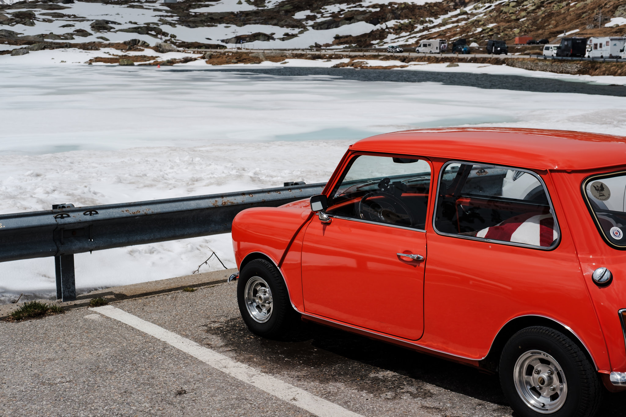 Red classic car parked on a road next to an icy lake in a mountainous landscape.