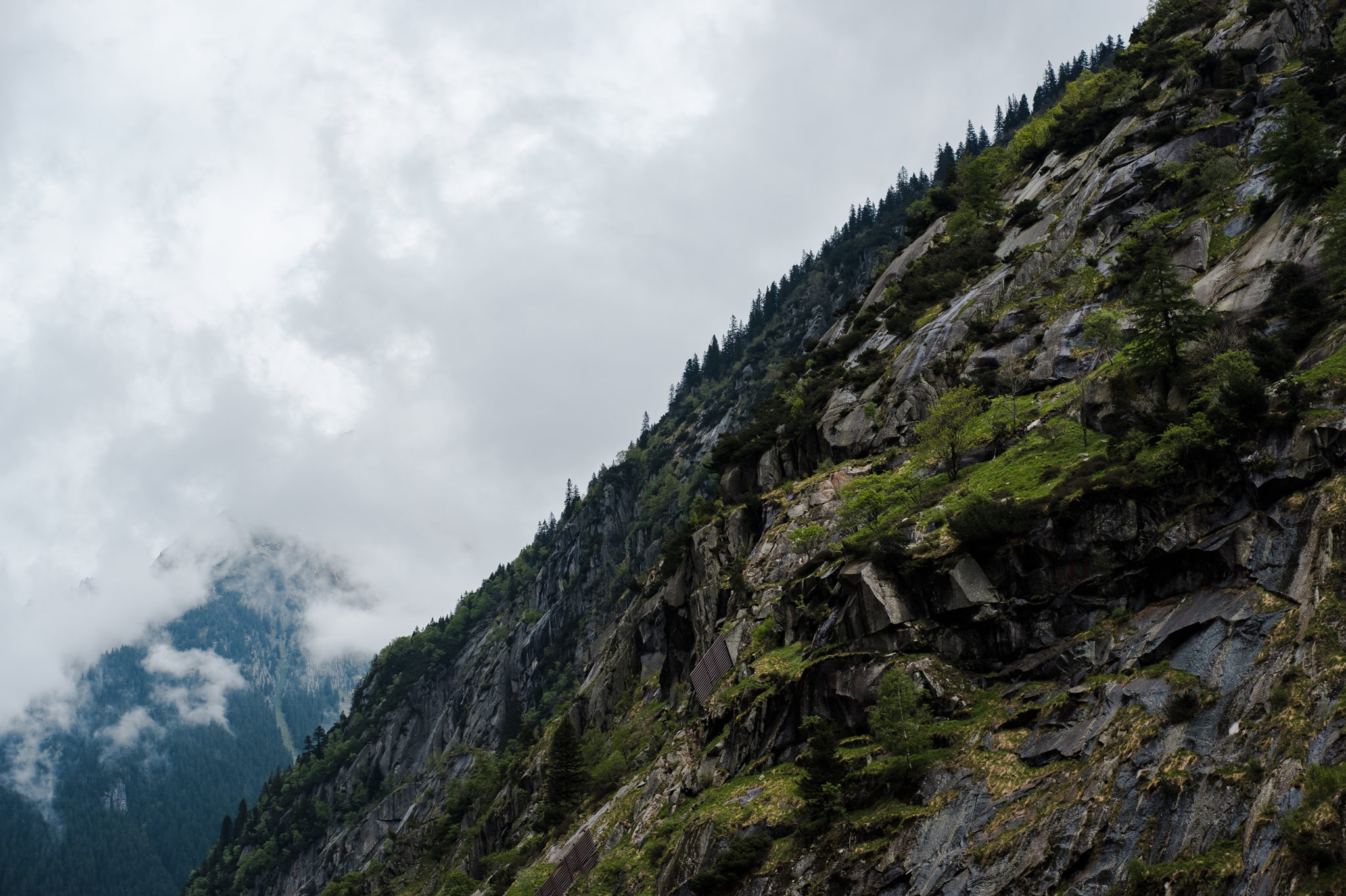 Mountain cliffside with vegetation, under an overcast sky. Clouds partially obscure another peak.