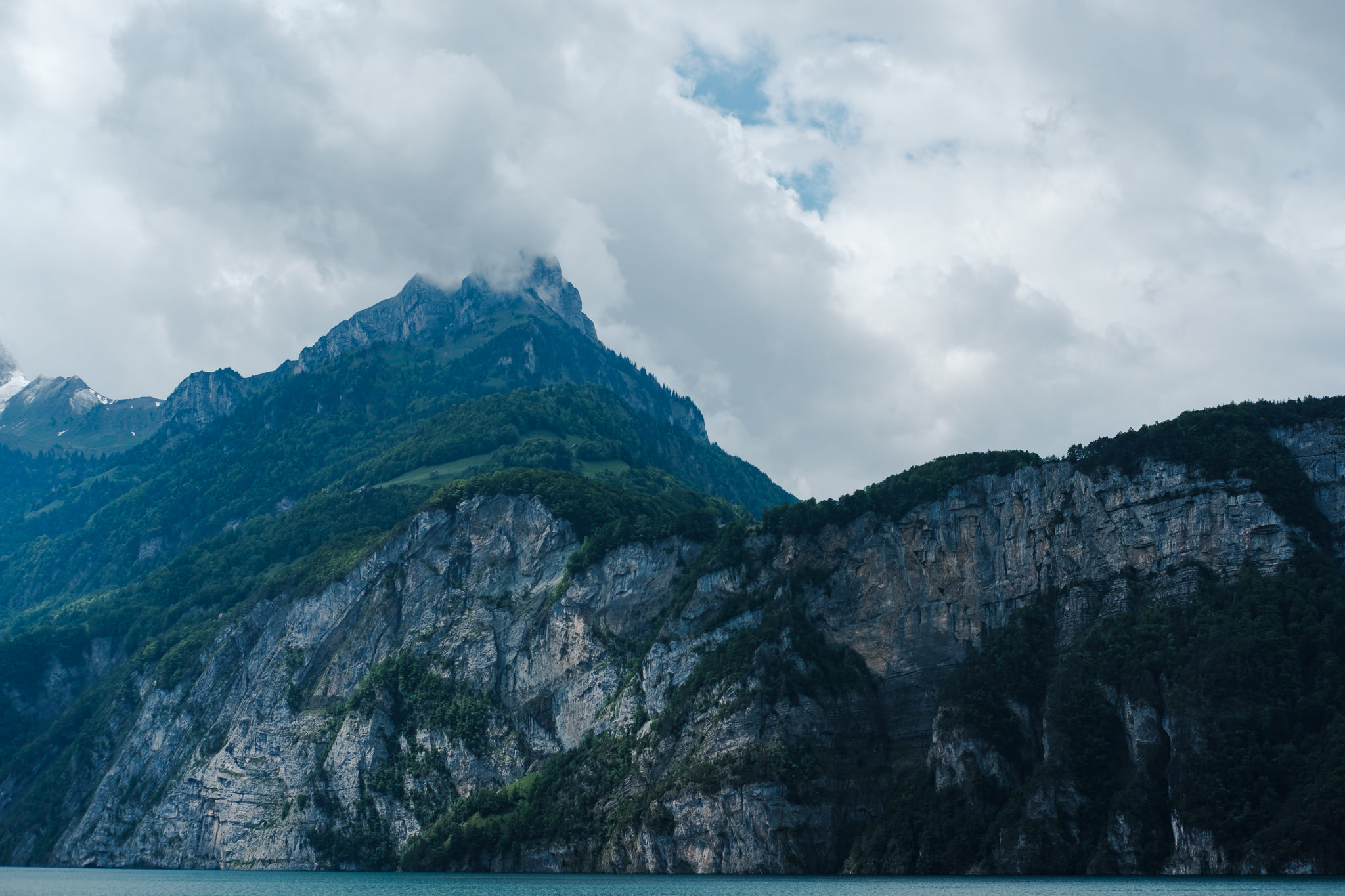 Mountain landscape with rocky cliffs, green trees, and cloudy sky. Water at the bottom.