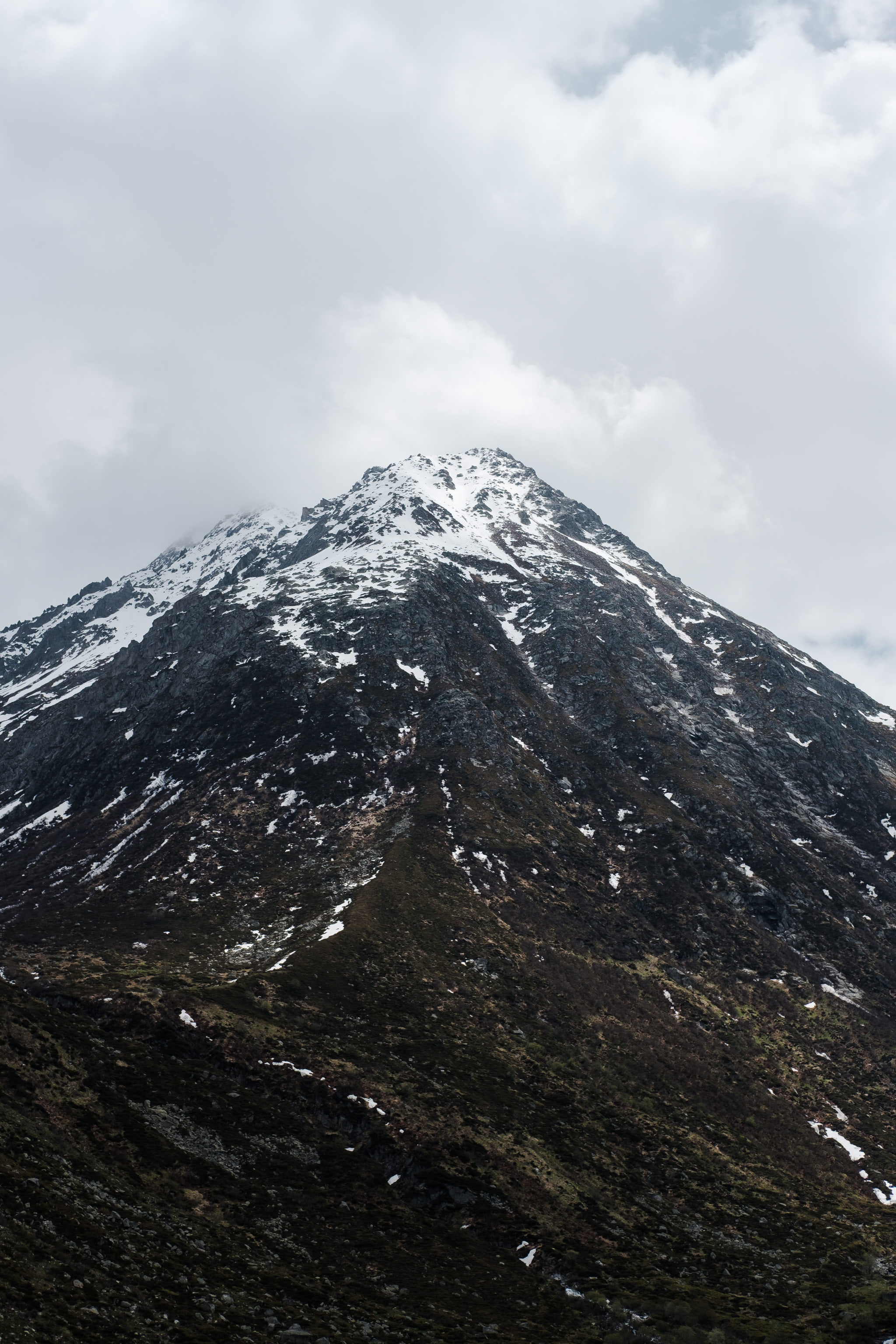 Mountain peak with snow and clouds. Dark rocks and green vegetation are visible. Cloudy sky.