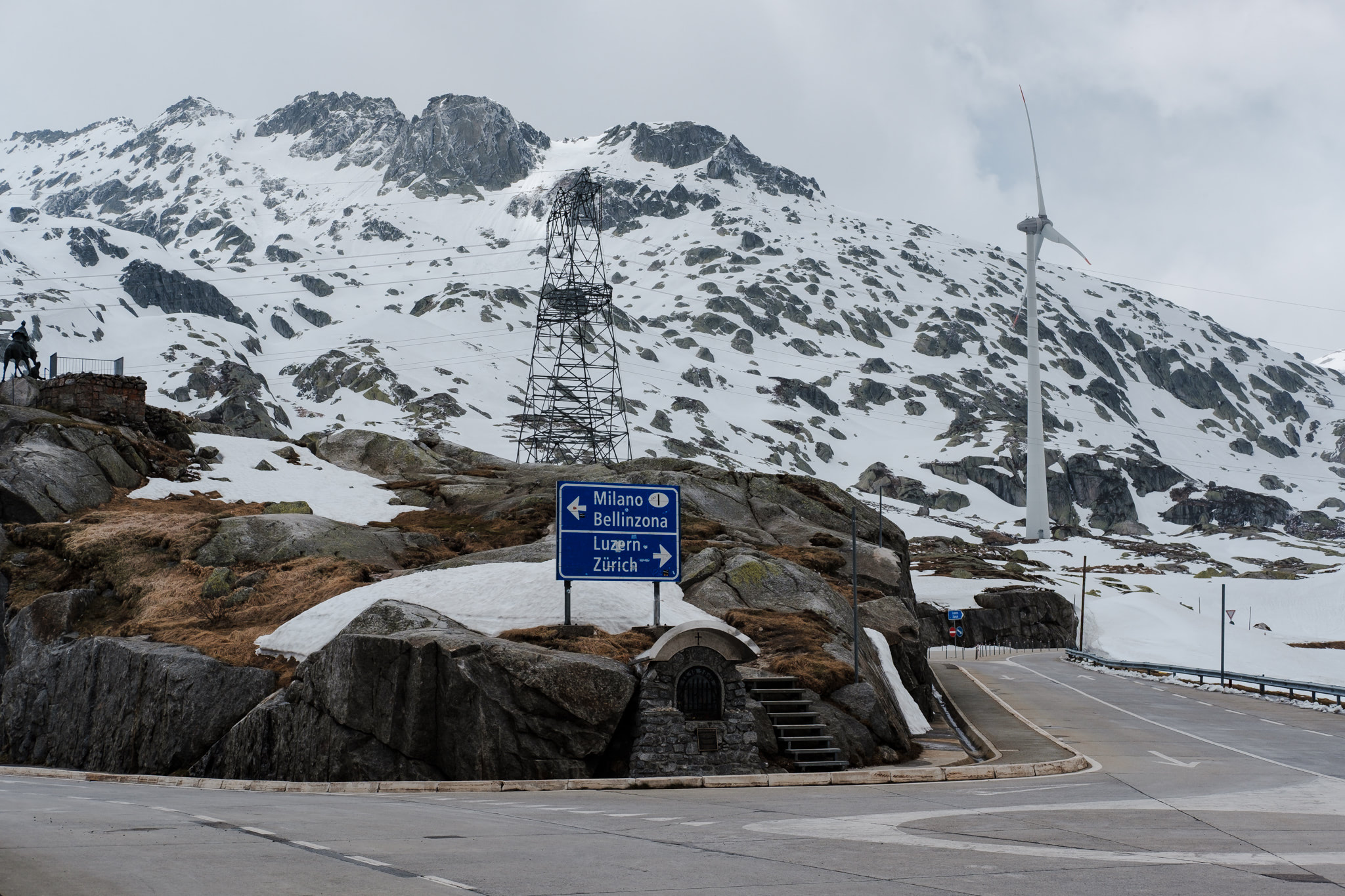 Road sign in mountainous landscape with snow. Wind turbine and power lines are visible.