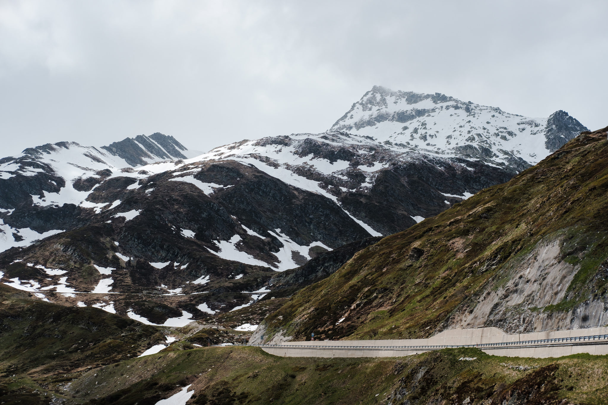 Mountain range with snowy peaks, road winding along the hillside. Overcast sky.