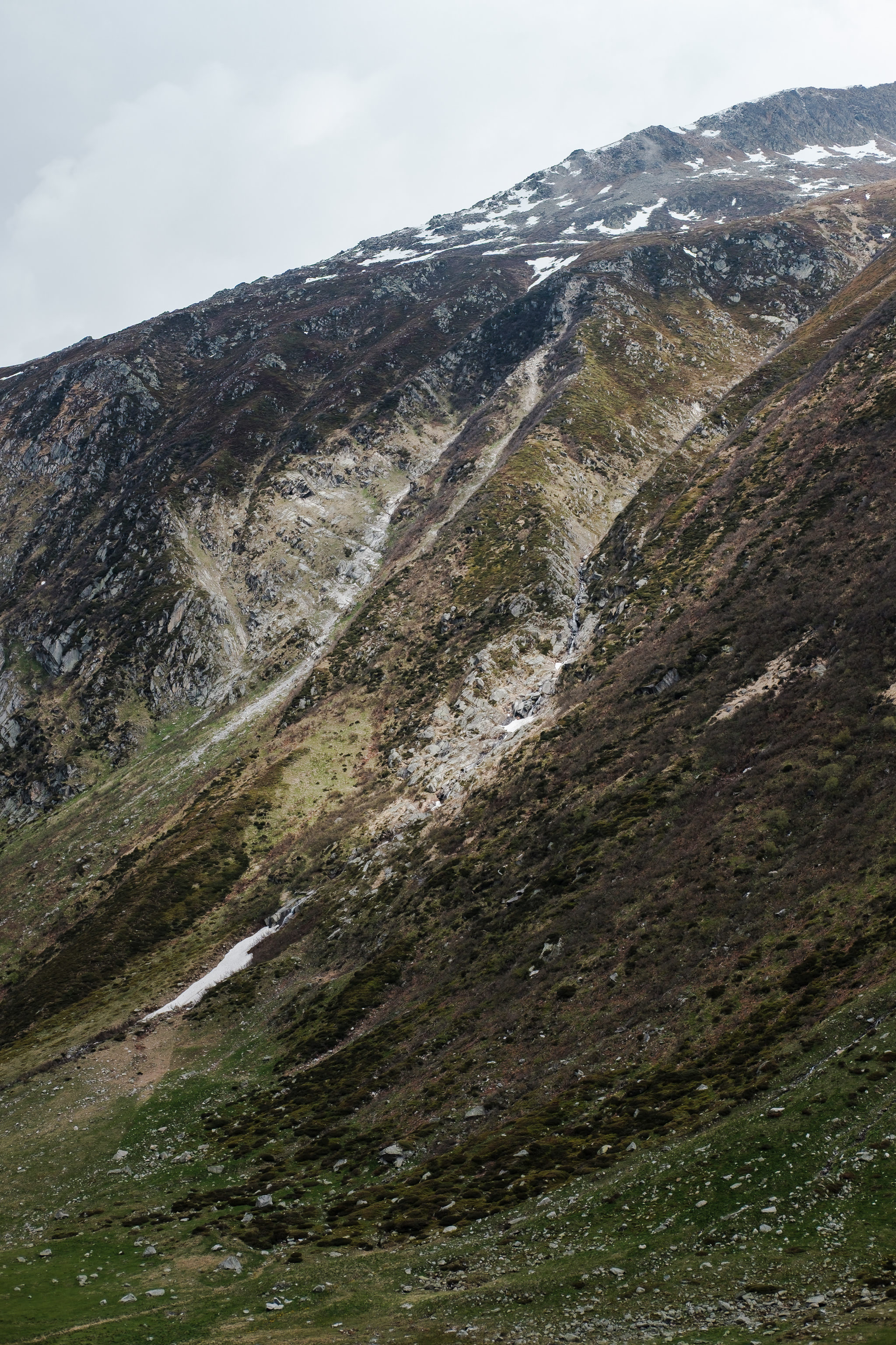 Mountain slope with snow, vegetation, and a stream.