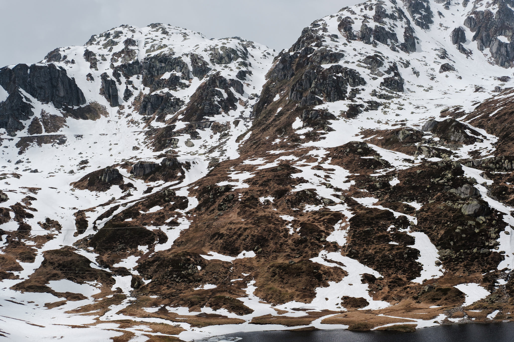 Snow-covered mountain landscape with exposed rock faces and a small body of water at the base.