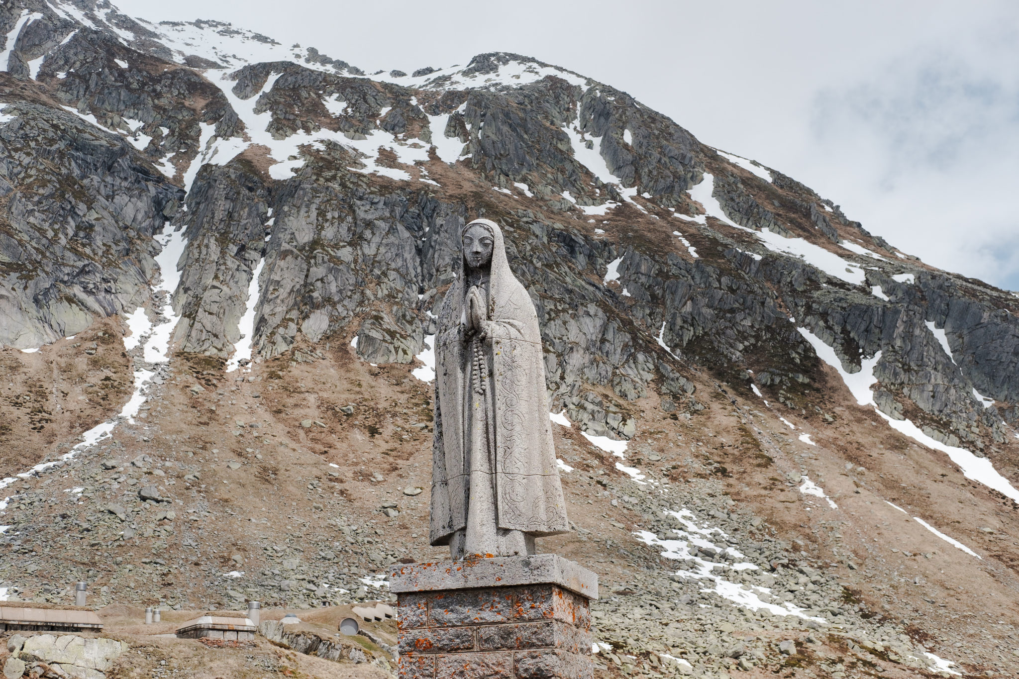 Statue of a hooded figure praying, mountains in the background with snow.
