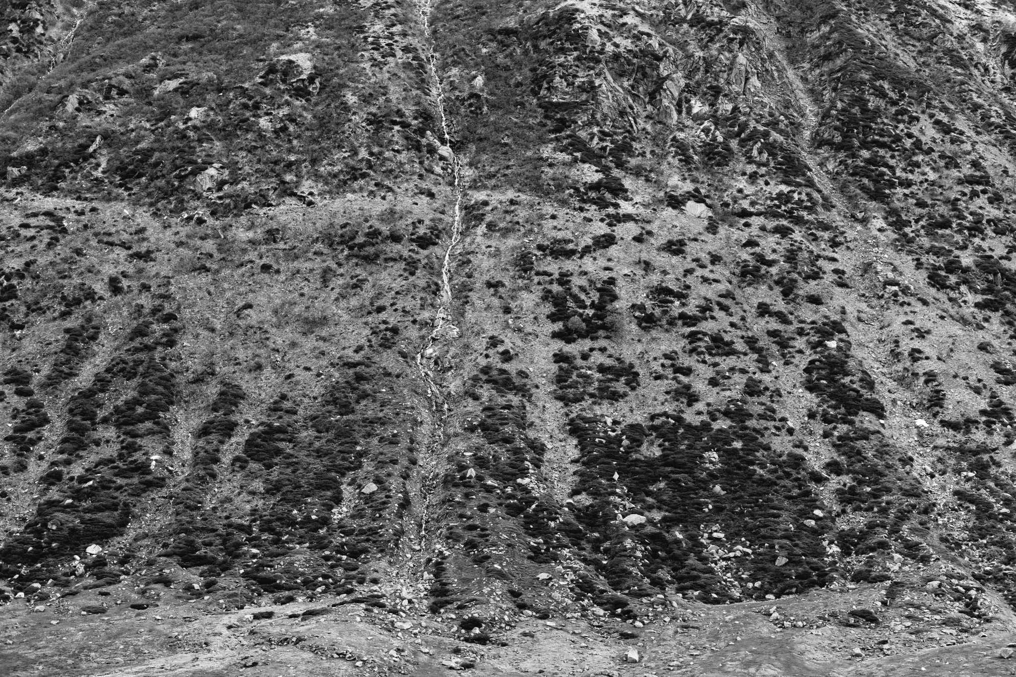 Black and white image of a mountain landscape. A waterfall cascades down the mountainside. The terrain is rocky and covered with vegetation.