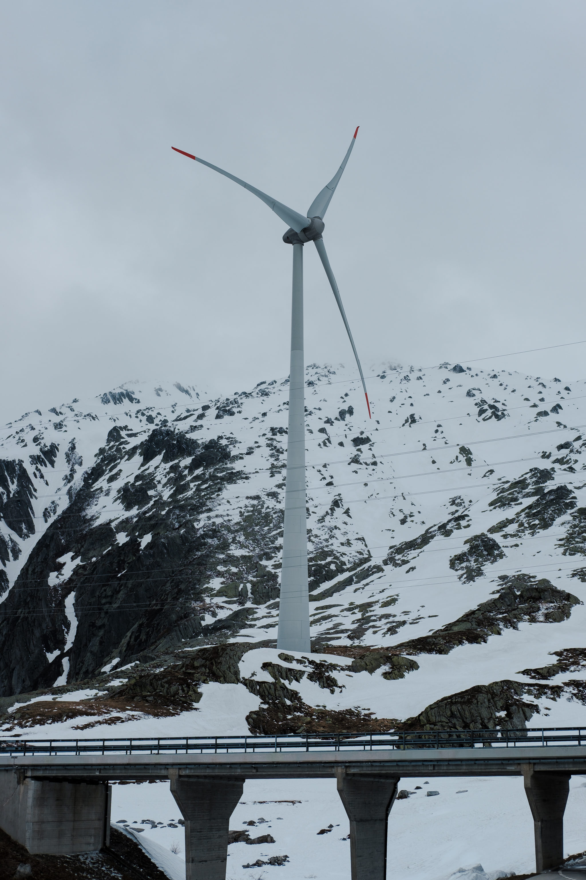 Wind turbine in a snowy mountain landscape, with a bridge in the foreground.