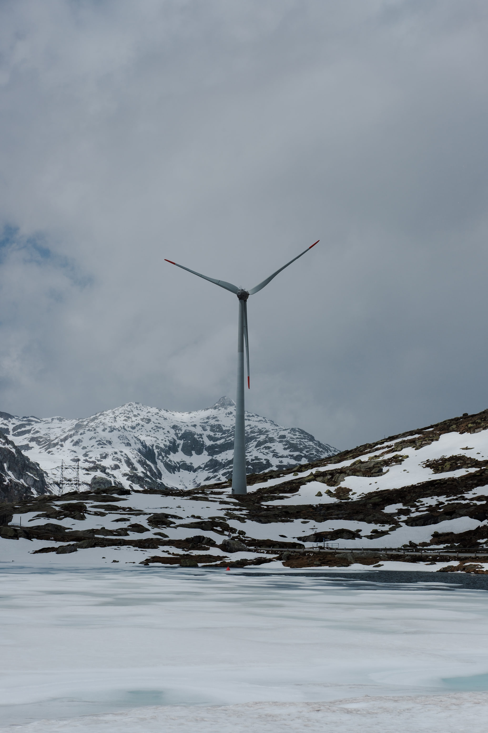 Wind turbine in a snowy mountain landscape. Icy lake in the foreground, cloudy sky.