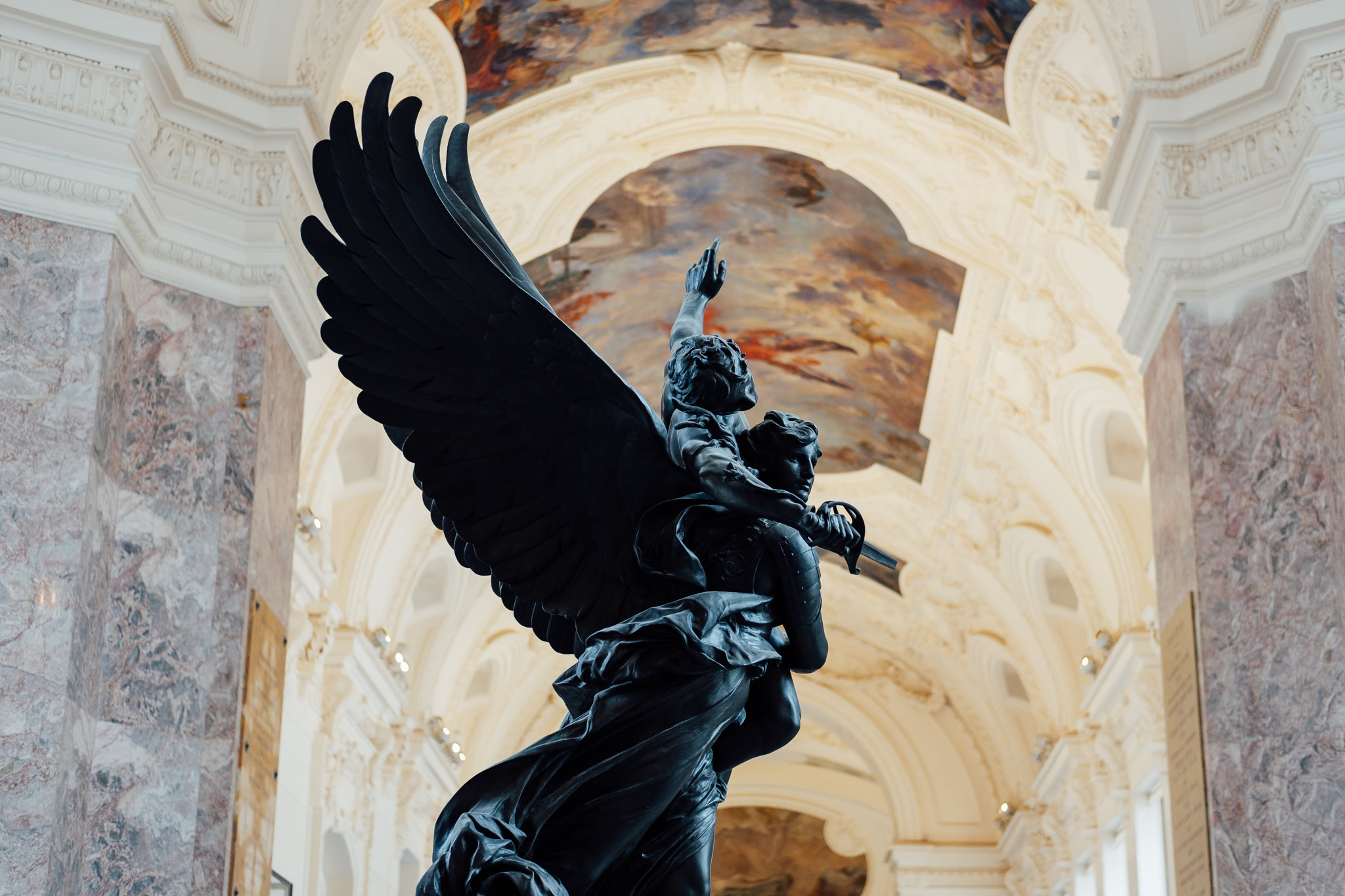 Dark statue of an angel with large wings in a grand interior with an ornate ceiling.