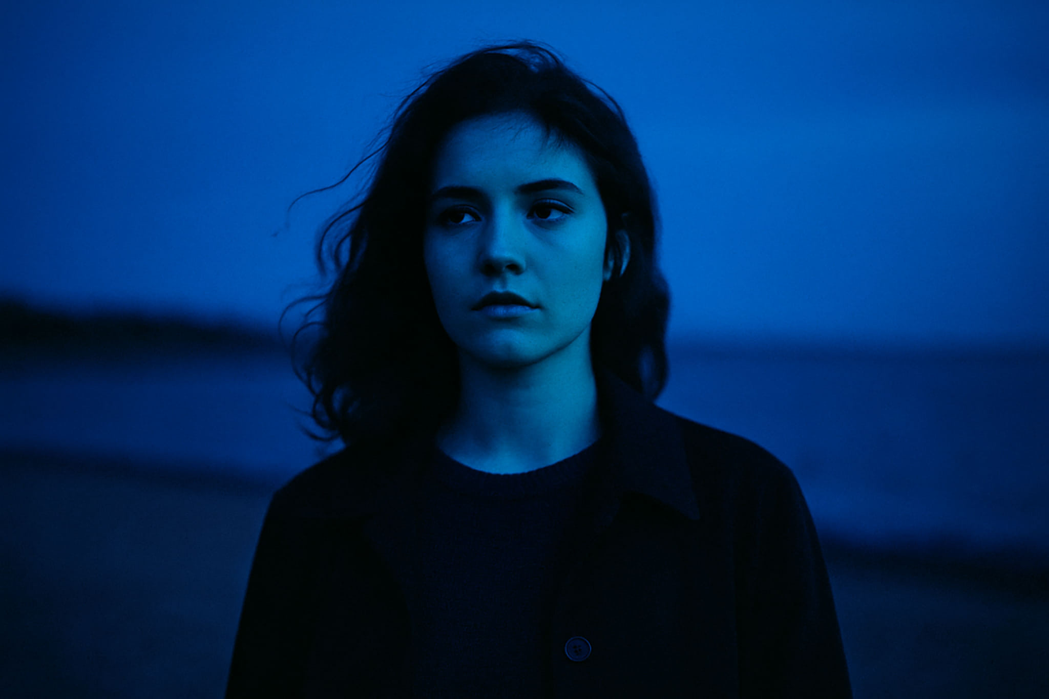 Woman with dark hair and coat, looking pensively towards the left, in blue dusk light.