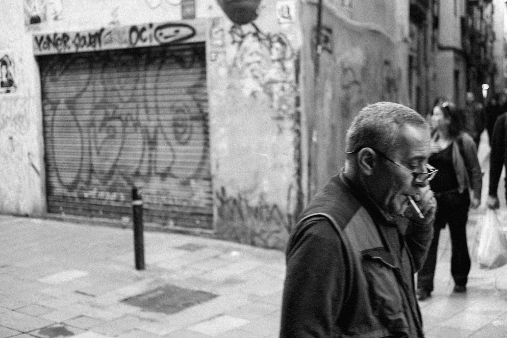 Black and white street photograph of a middle-aged man smoking a cigarette while walking past a building with graffiti-covered, closed metal shutters.