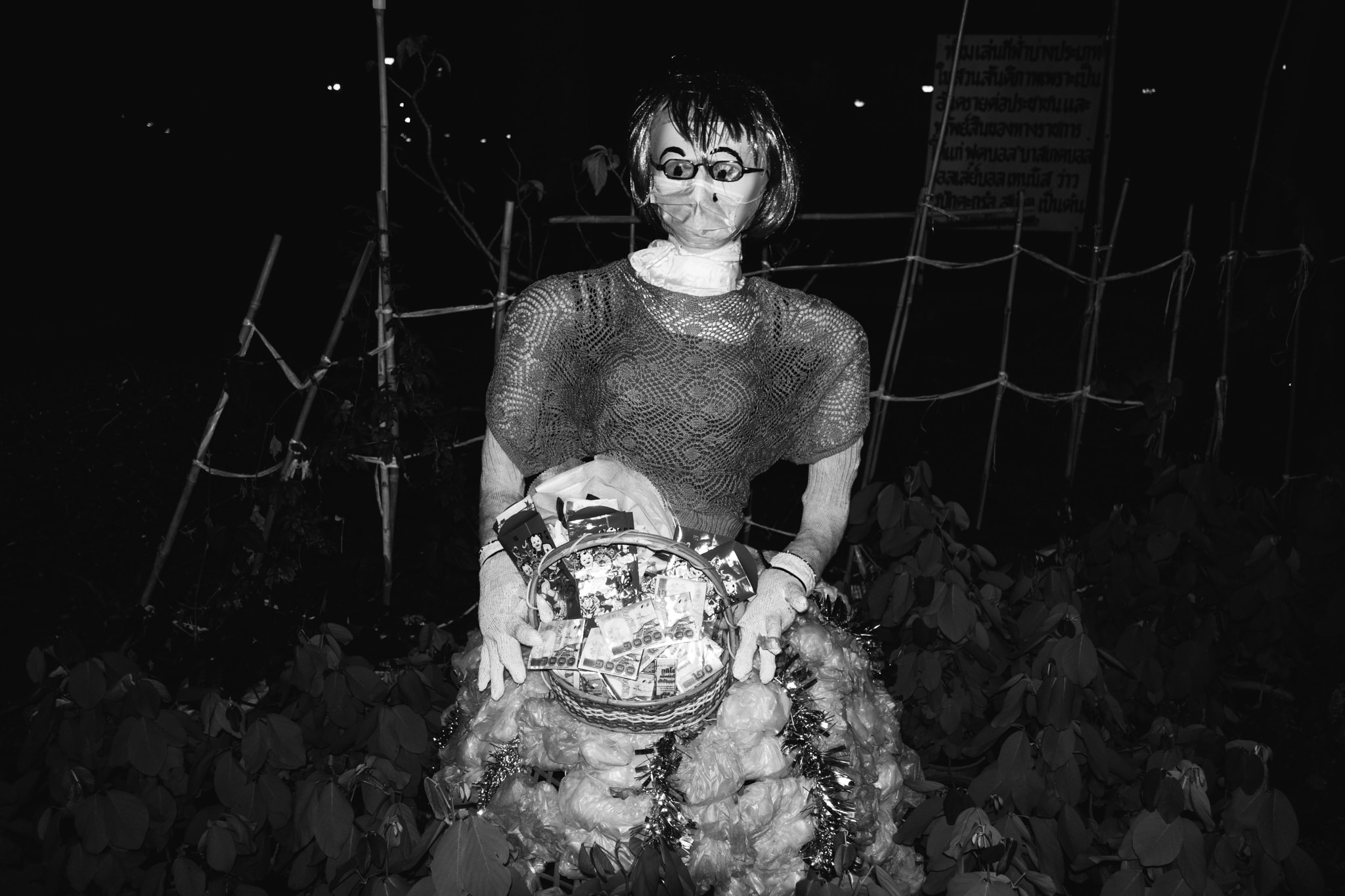 Black and white photo of a disturbing effigy or scarecrow at night, featuring a plastic mask with glasses and a medical mask, holding a basket full of money, set against a dark, leafy background with bamboo structures.