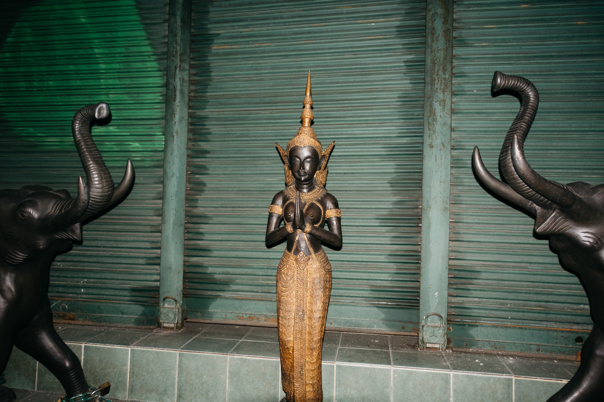 Dark bronze statue of a female figure in traditional Thai dress with hands in a prayer position, flanked by two dark elephant statues, set against a teal corrugated metal background.