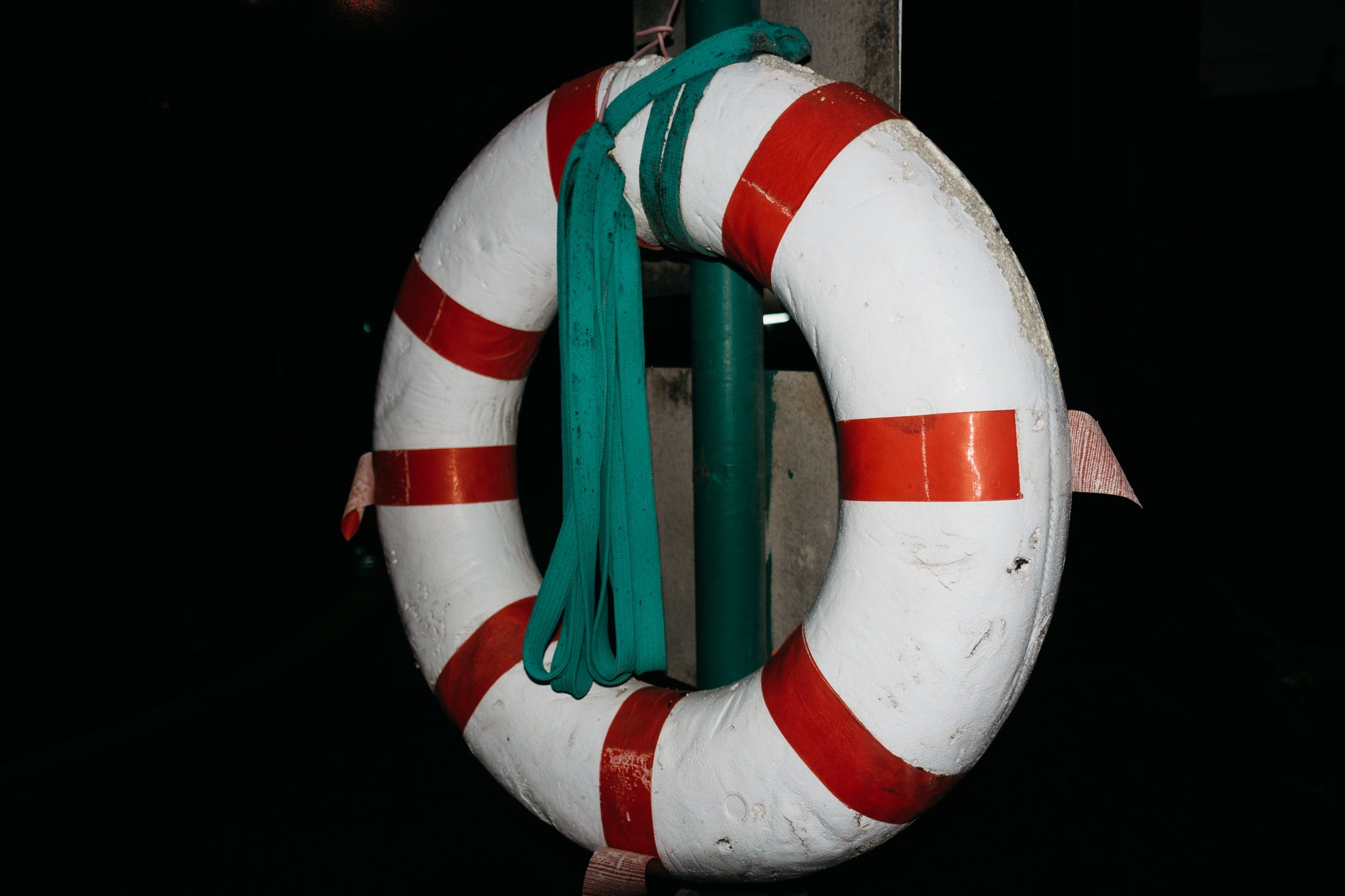 Close-up of a worn white life preserver ring with thick red reflective stripes, hanging against a dark background with a green pole.
