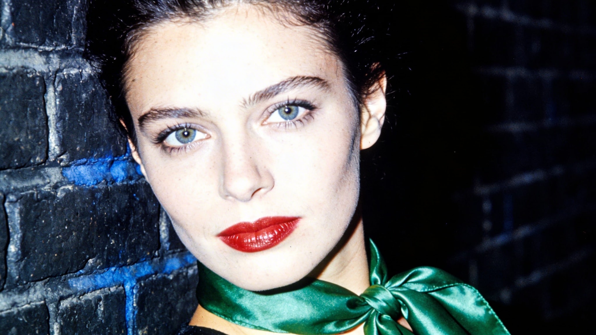 A woman with red lipstick and a green silk scarf leans against a dark brick wall in a grainy, high-contrast 1970s film portrait.