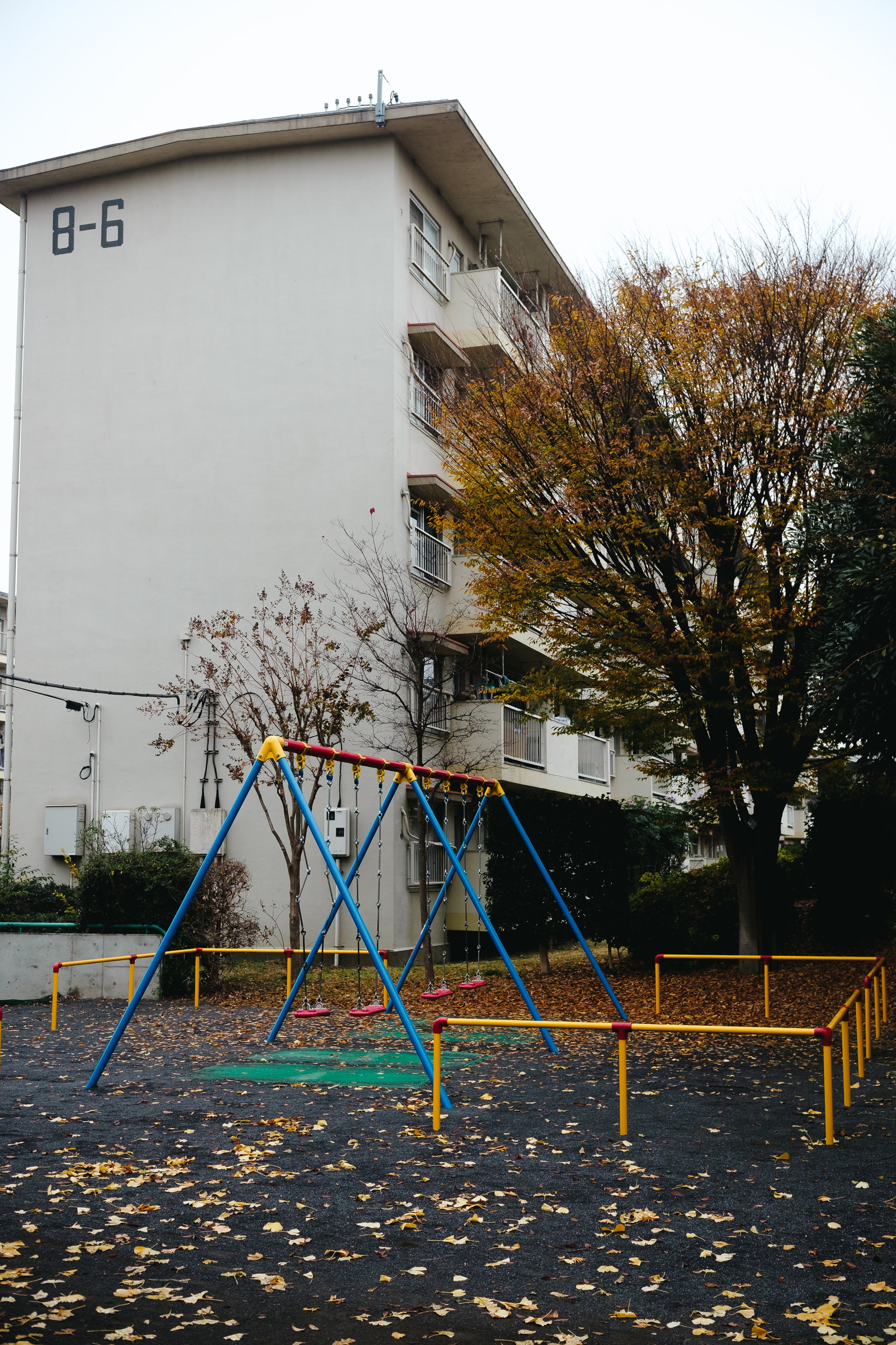 Empty playground with a blue and yellow swing set in front of a white residential apartment building during autumn.