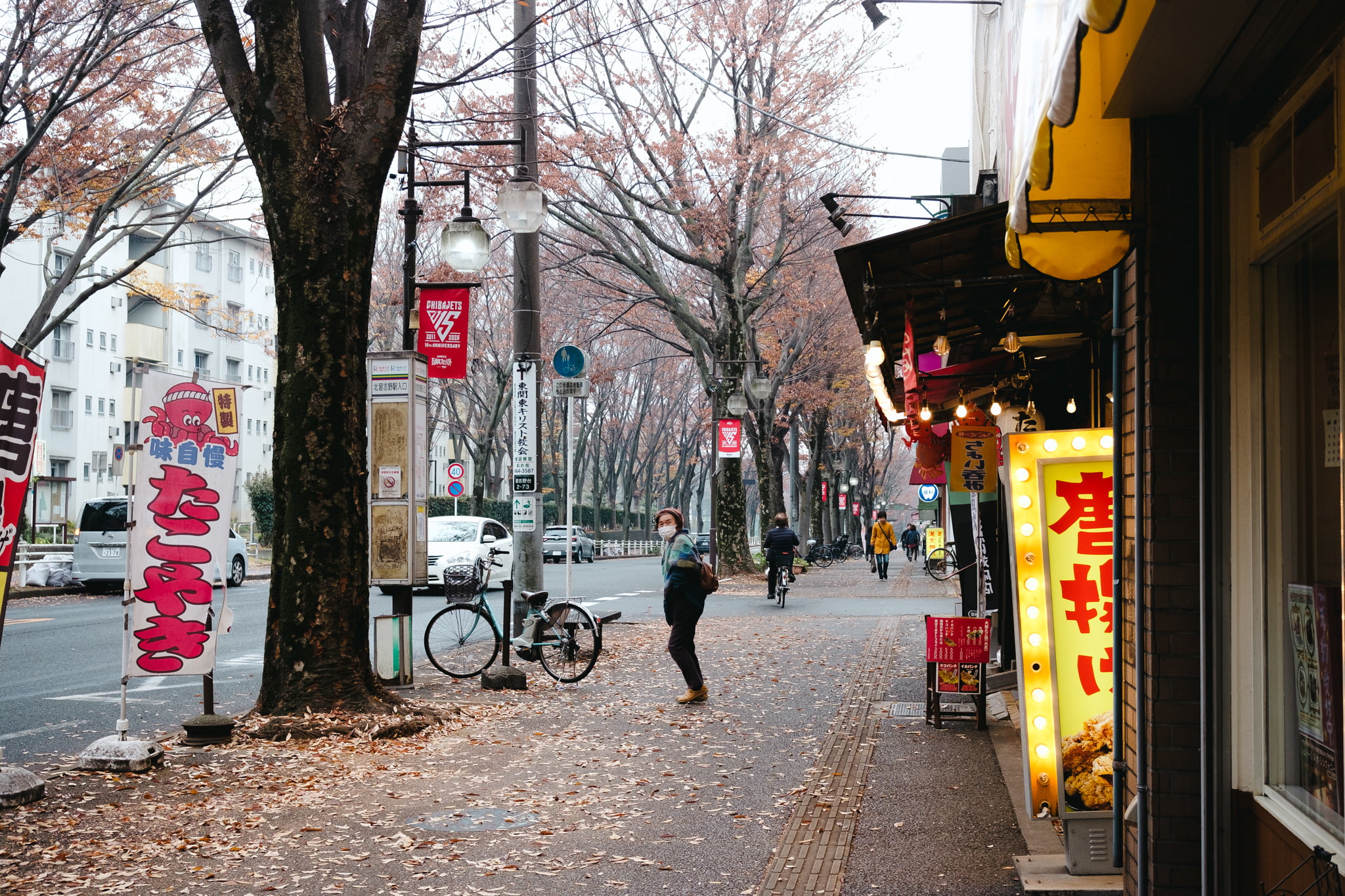 A woman wearing a face mask walks along a sidewalk lined with trees and small shops in Japan during autumn.