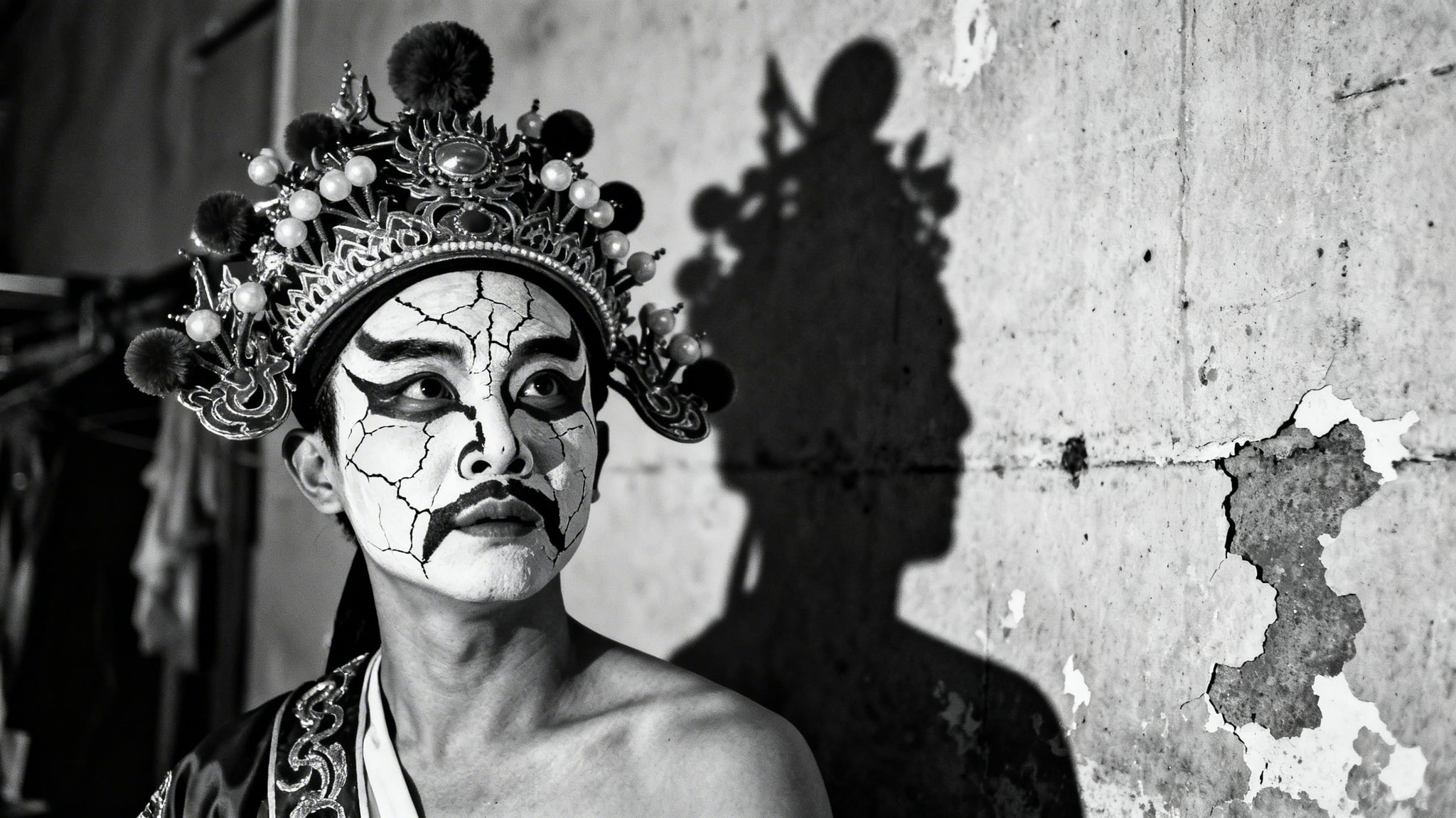 Black and white candid photograph of a Sichuan Opera performer backstage with cracked face paint and a dramatic shadow.